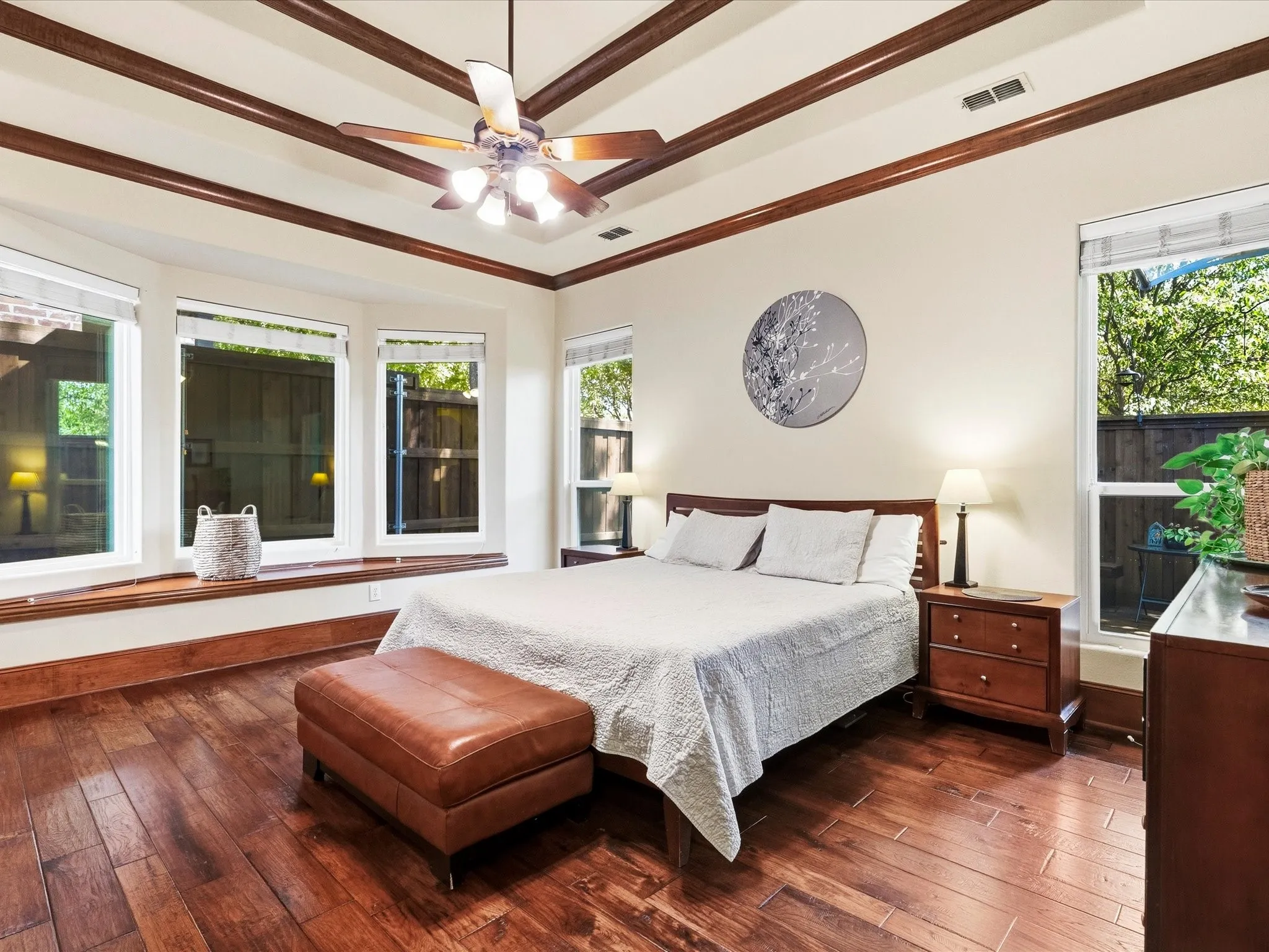 Wood-finish flooring throughout featuring a bay window with a built-in window seat, tray ceiling with decorative wood beams, and a ceiling fan with integrated lighting