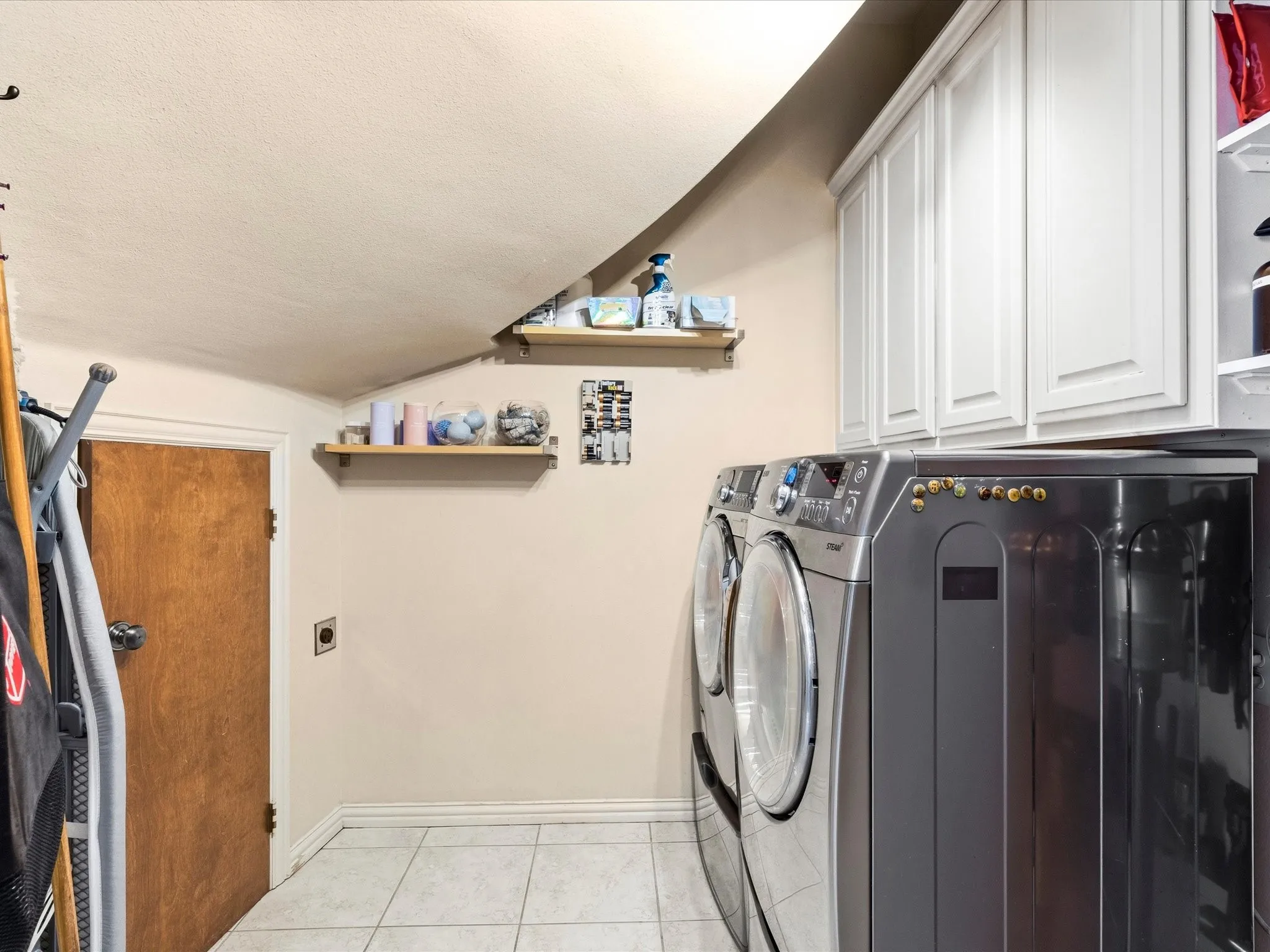 Laundry area featuring extra storage space, upper cabinetry, two wall-mounted shelves, and light-toned tile flooring