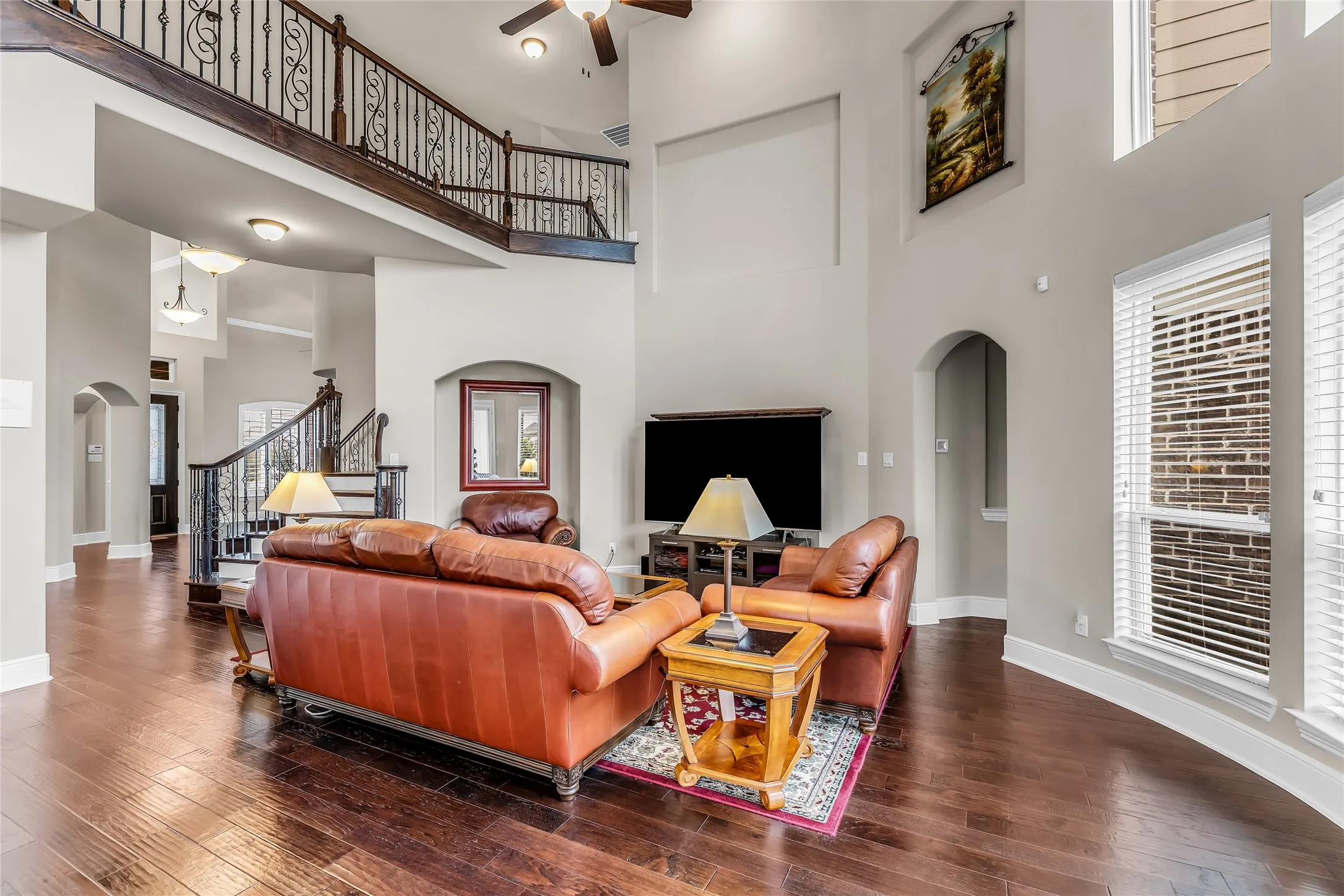 Spacious living area featuring high ceilings, wood-finish flooring, a grand staircase with wrought iron balusters, and multiple large windows with blinds