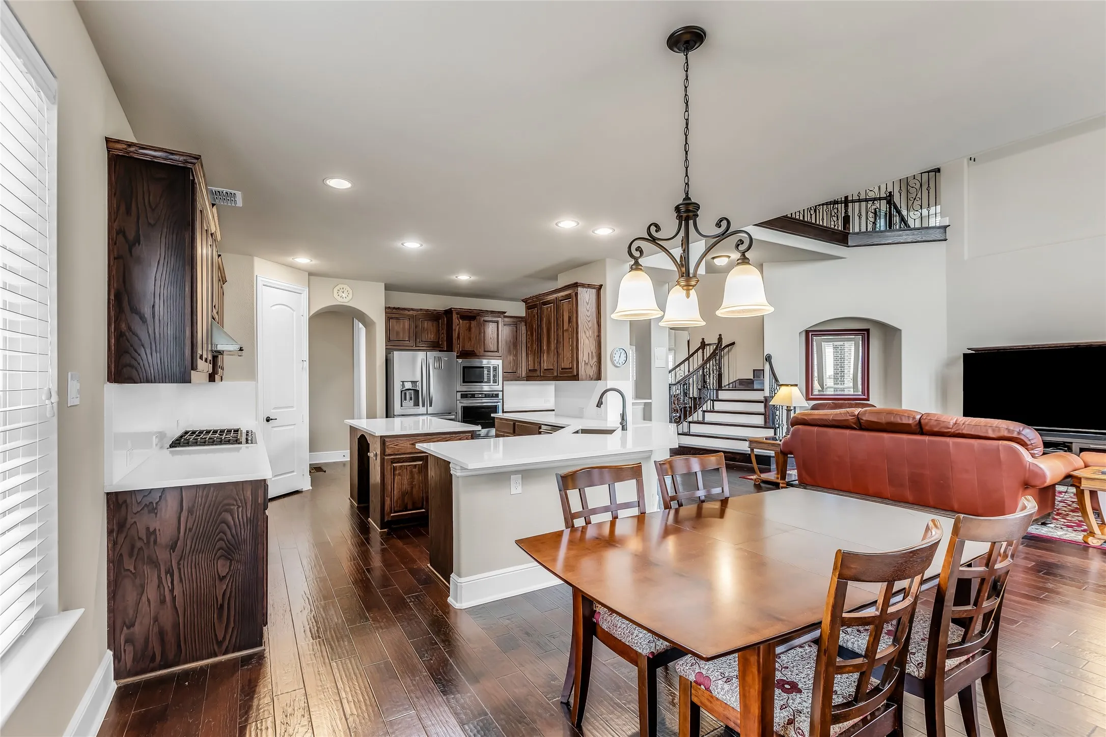 Open-concept living area featuring rich wood-finish flooring, a kitchen with stainless steel appliances and white countertops, a dining area with a chandelier, and a prominent staircase with wrought iron balusters