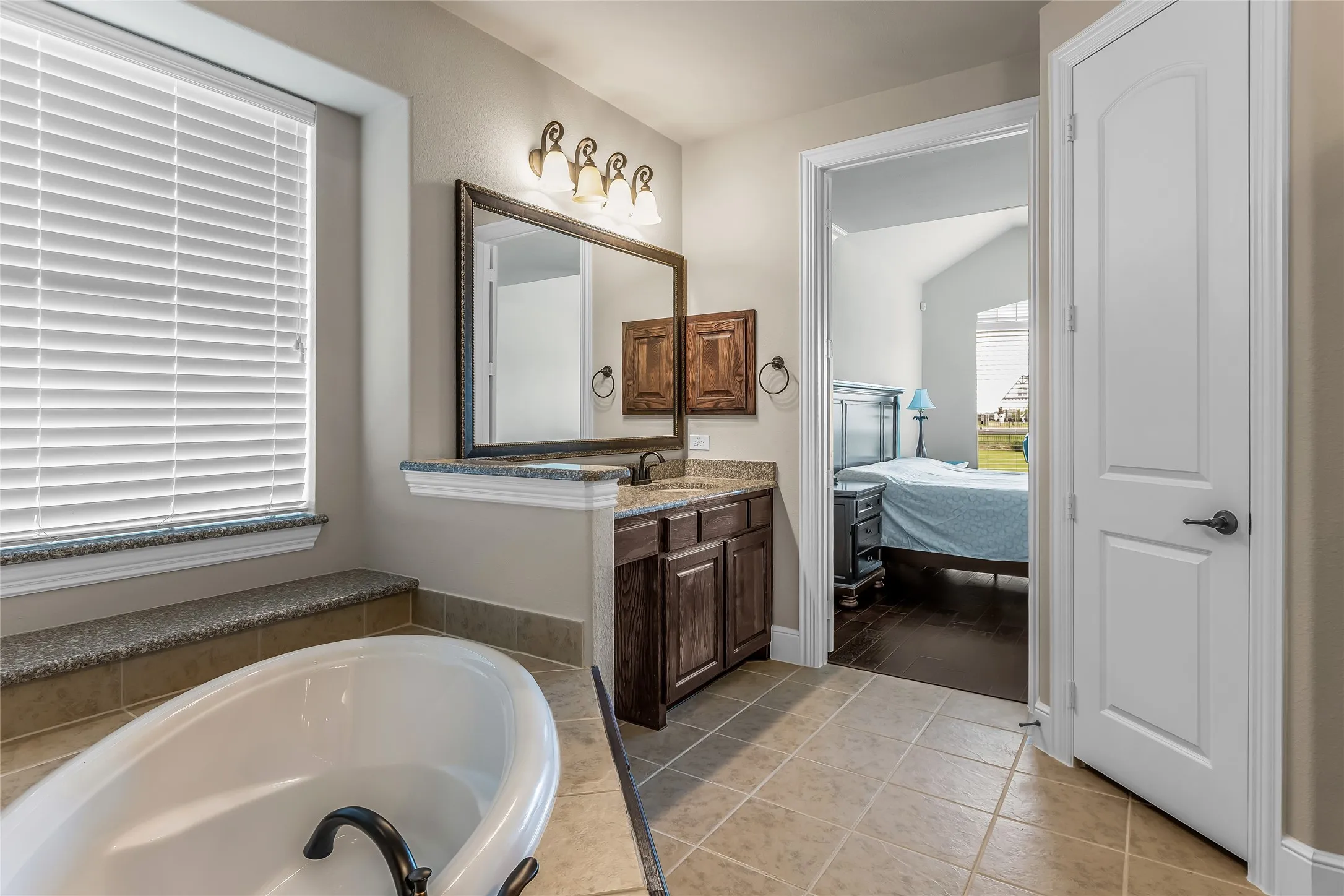 Freestanding soaking tub with dark bronze fixtures, tile flooring, wood-finish vanity with stone countertop, framed mirror, and a window with horizontal blinds