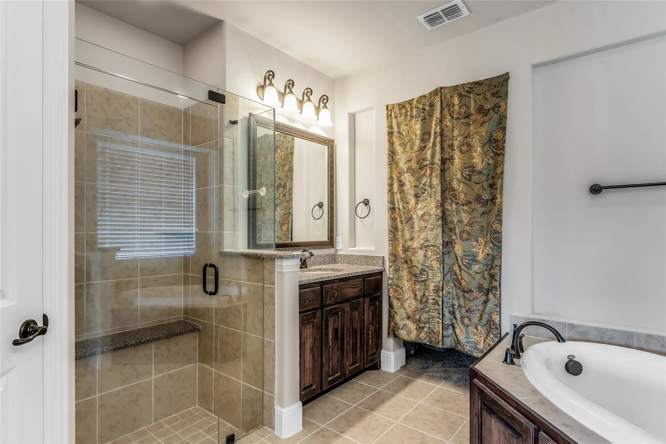 Bathroom featuring a glass-enclosed shower with a built-in bench, a separate soaking tub, a vanity with wood-finish cabinetry and a stone countertop, and tile flooring