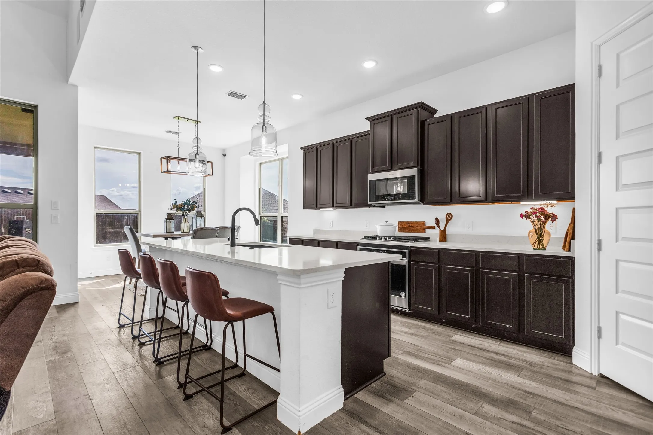 Kitchen with light wood finished floors, hanging light fixtures, a kitchen breakfast bar, dark brown cabinetry, and a kitchen island with sink