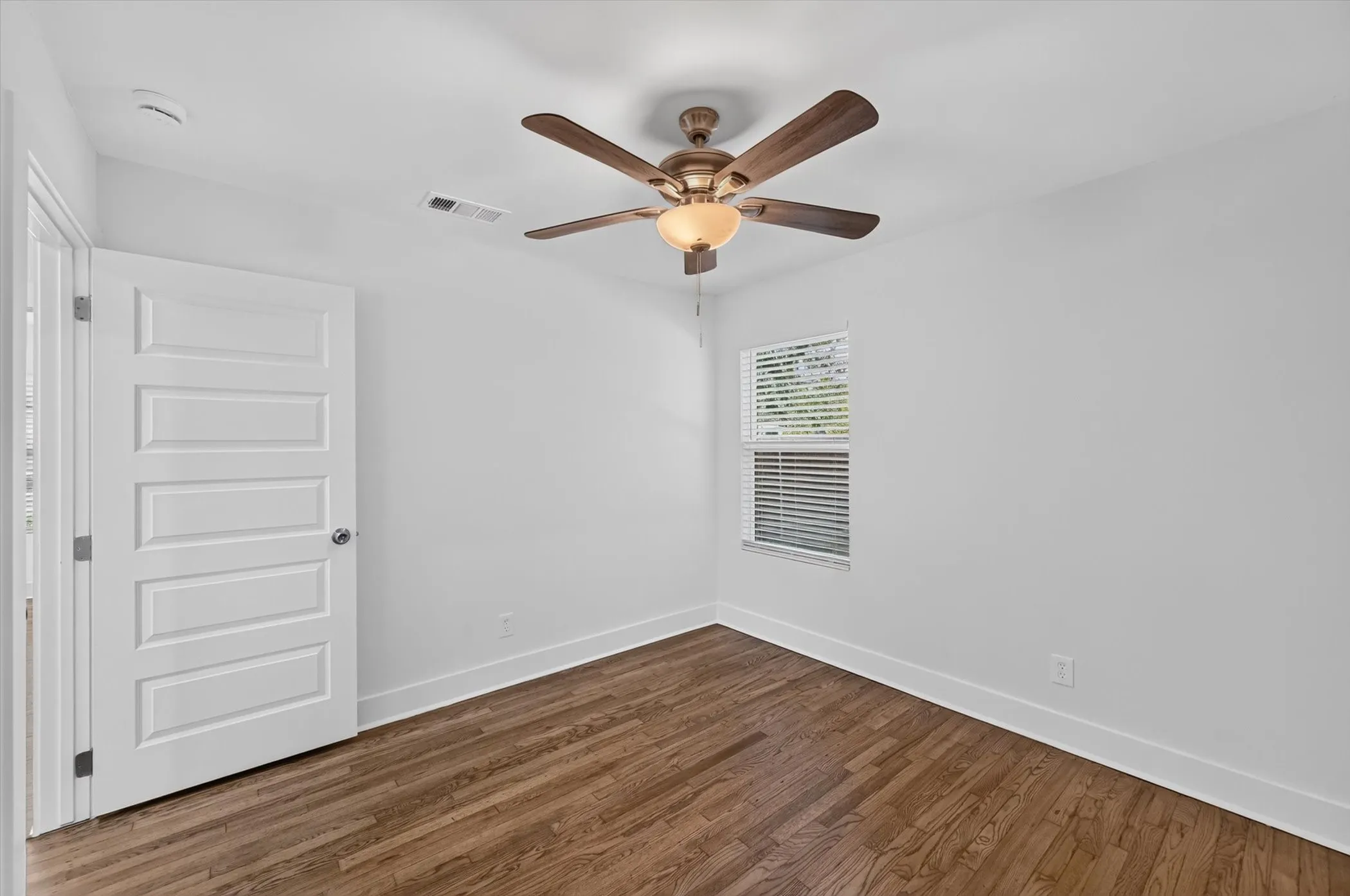 Bedroom featuring wood-finish flooring, a ceiling fan with integrated lighting, a single window with blinds, and a paneled interior door