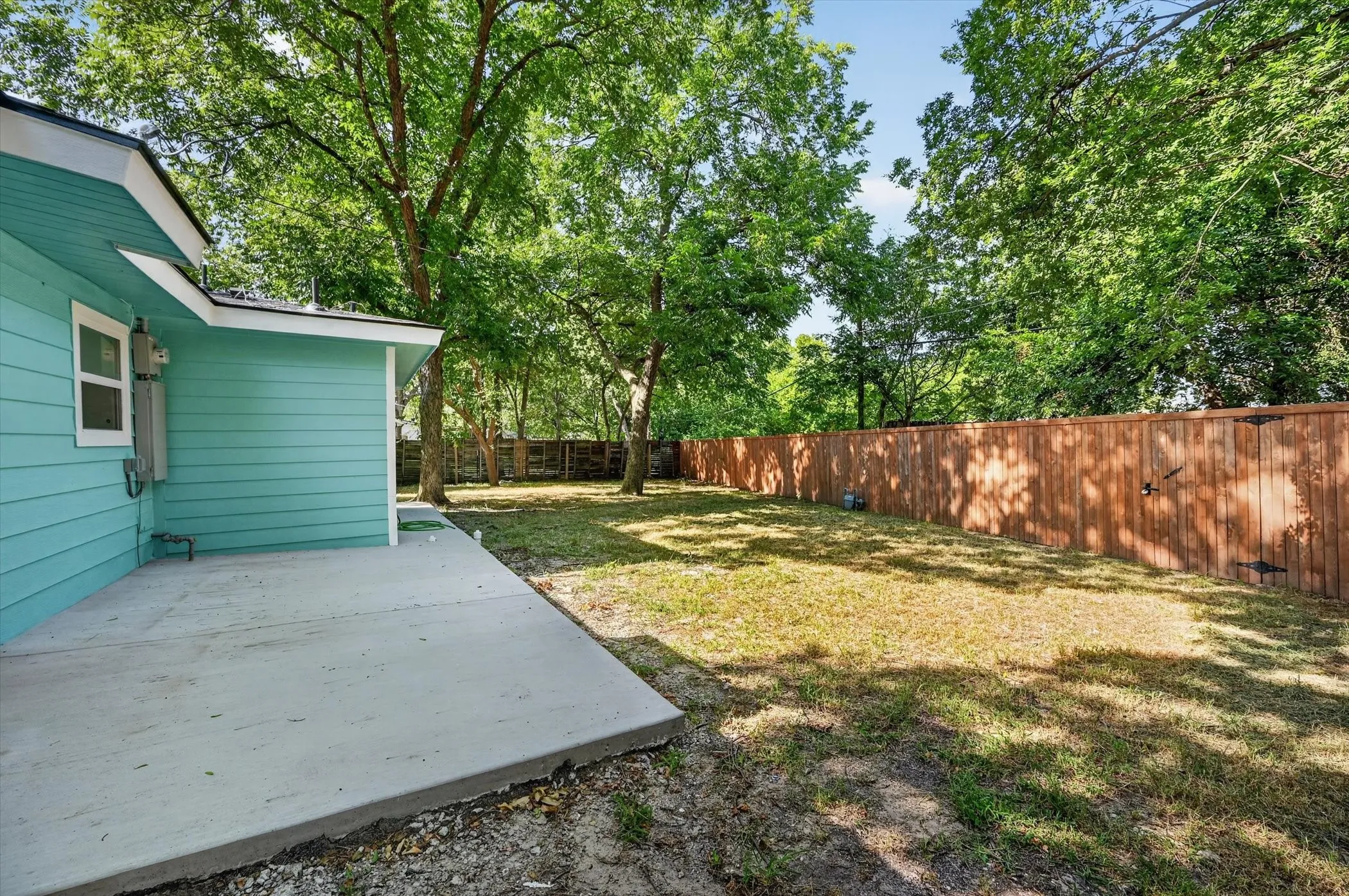 Expansive backyard featuring a concrete patio, mature trees, and a tall wood fence