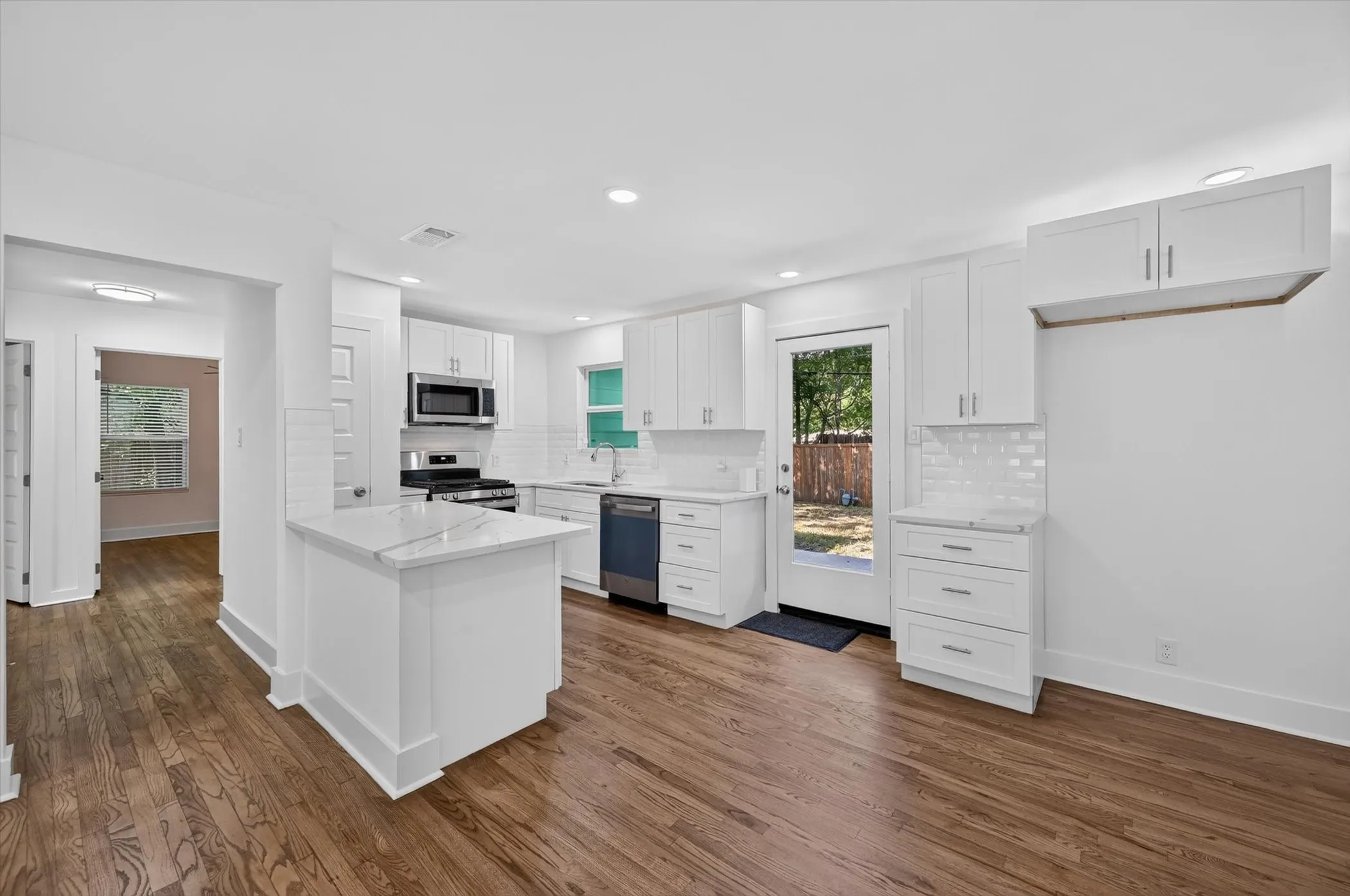 Contemporary kitchen featuring a central island with a stone countertop, white shaker-style cabinetry, subway tile backsplash, stainless steel appliances, and wood-finish flooring