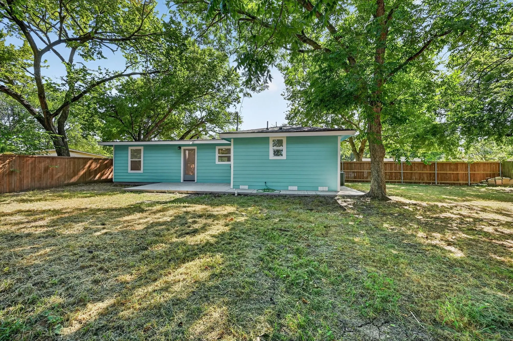Expansive backyard featuring a teal-sided home with white trim windows, a concrete patio, and a wood privacy fence