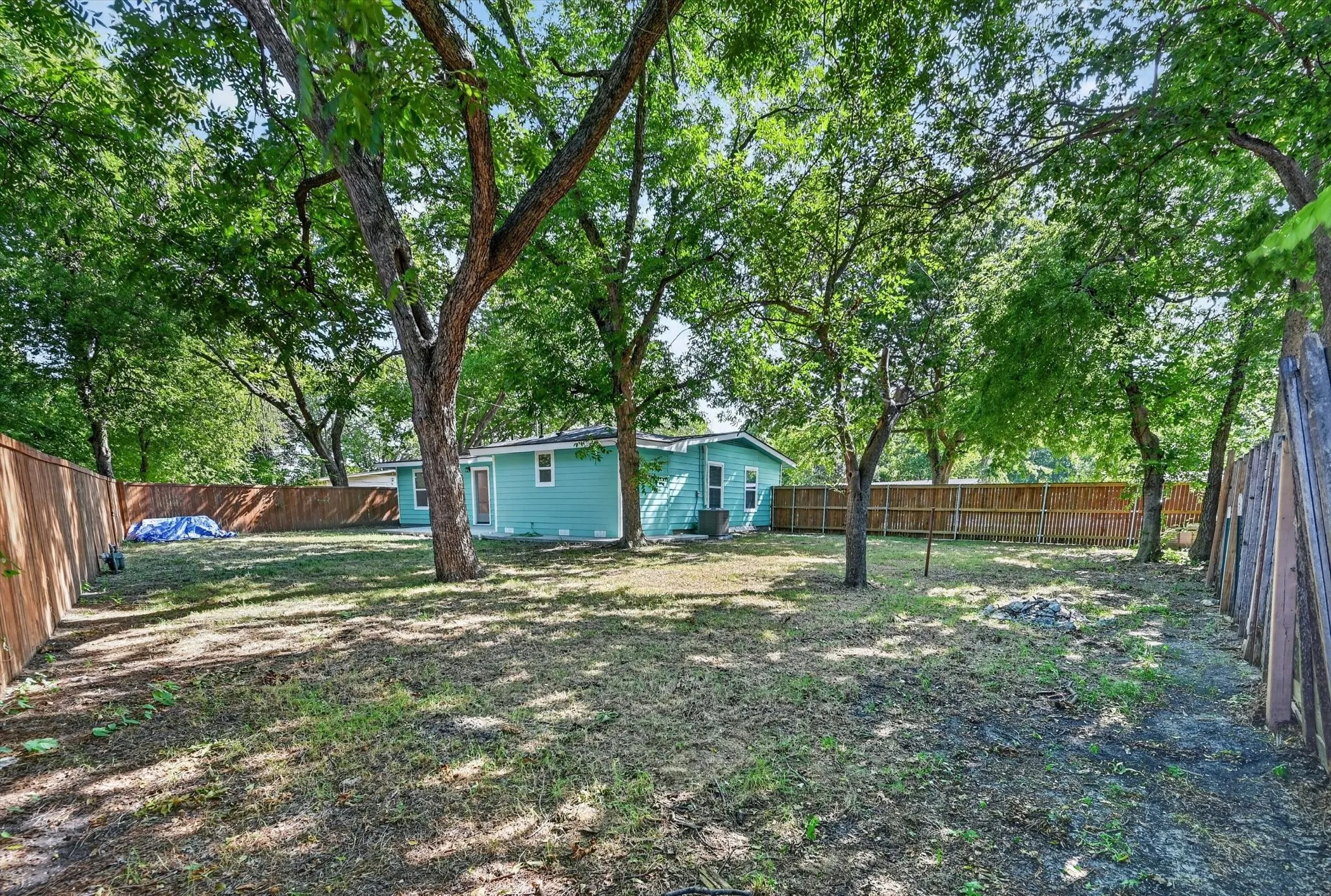 Expansive backyard featuring mature trees providing ample shade, a wood privacy fence, and a teal-colored siding structure