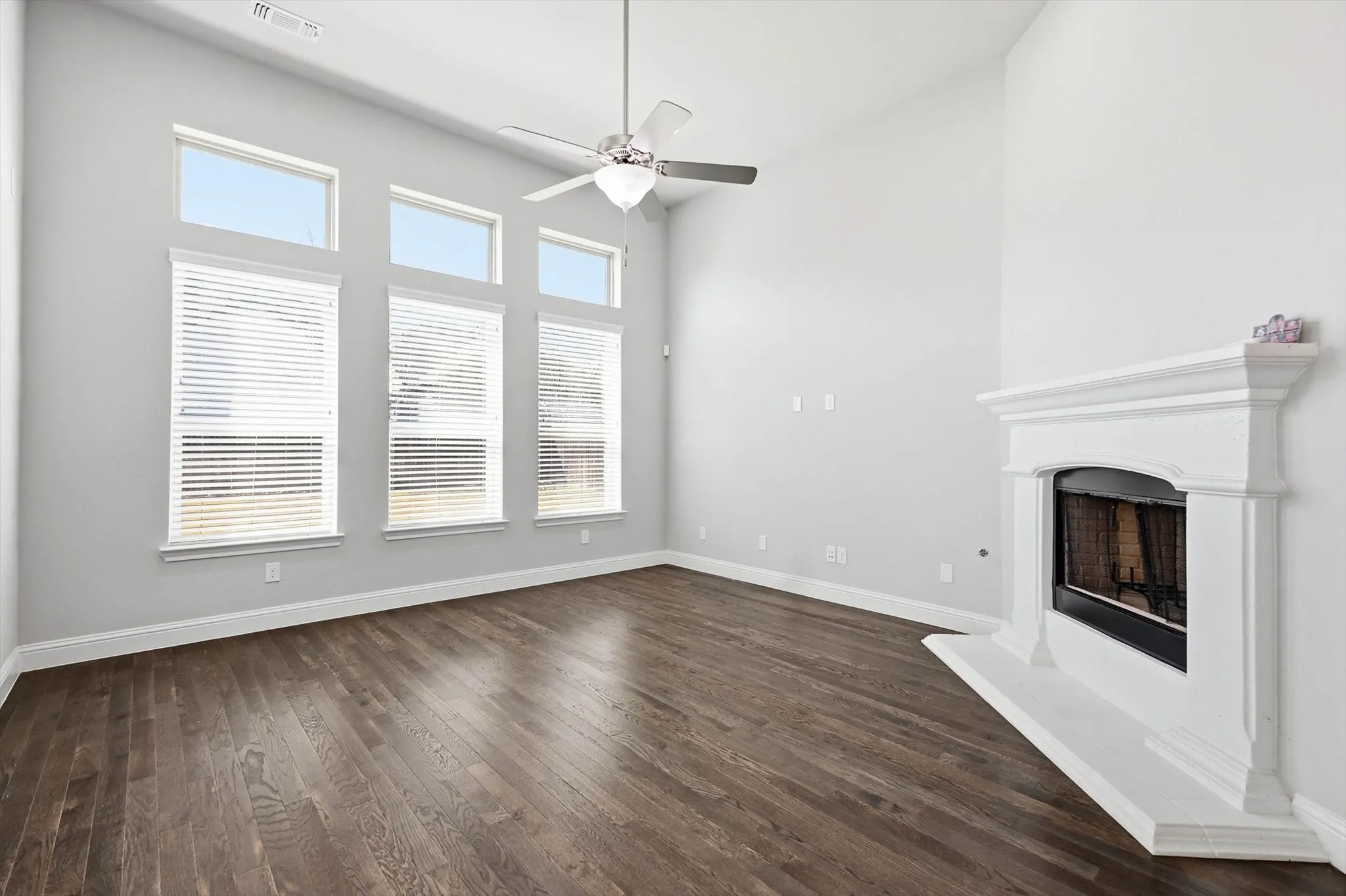 Spacious living area featuring high vaulted ceilings, multiple transom windows, wood-finish flooring, a ceiling fan fixture, and a white mantel fireplace with a hearth