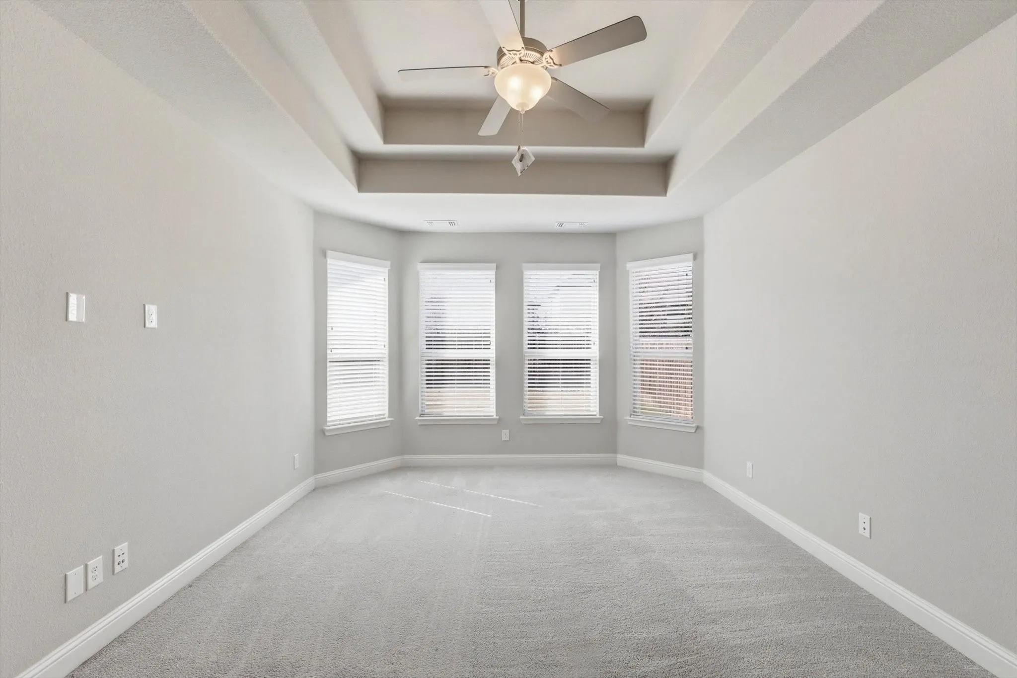 Spacious room featuring a tiered tray ceiling with a ceiling fan, light gray wall paint, and matching light gray carpeting