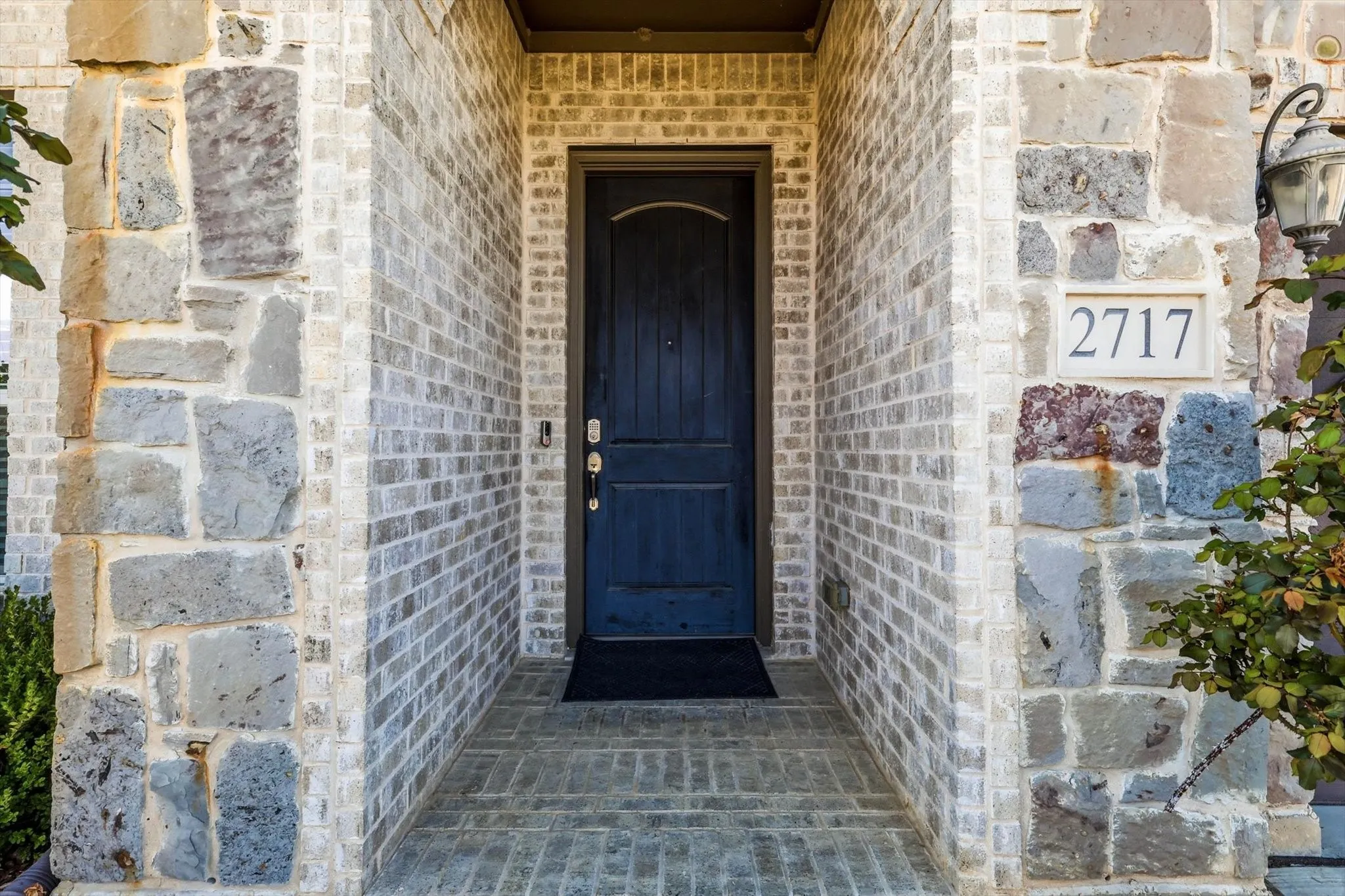 Architectural stone and brick facade with a dark paneled entry door, overhead soffit, brick paver entryway, and an exterior wall lantern