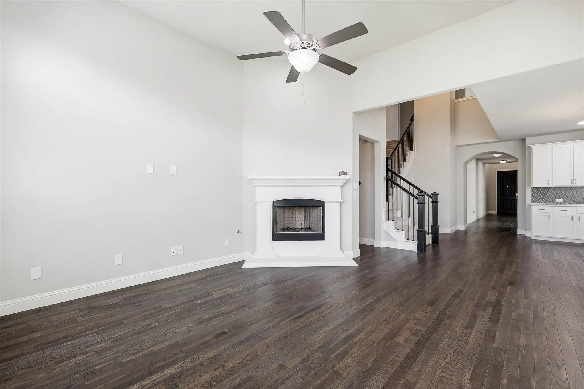 Open-concept living space with dark wood-finish flooring, a contemporary fireplace with a white mantel, and a ceiling fan