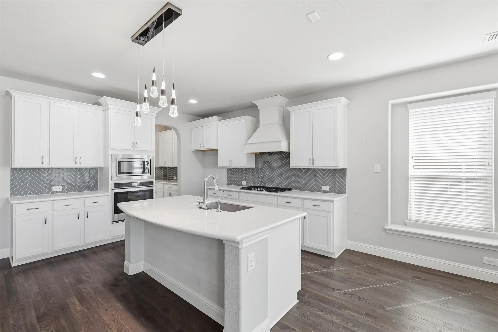 Kitchen featuring a large central island with a sink, white cabinetry, built-in stainless steel appliances, herringbone-patterned tile backsplash, and wood-finish flooring