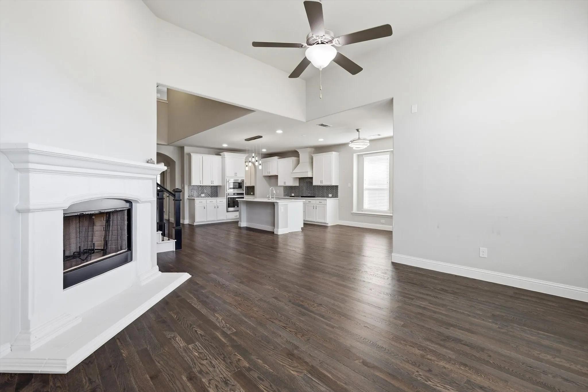 Spacious living area featuring a white mantel fireplace, wood-finish flooring, a vaulted ceiling with a ceiling fan, and an open concept layout connecting to the kitchen
