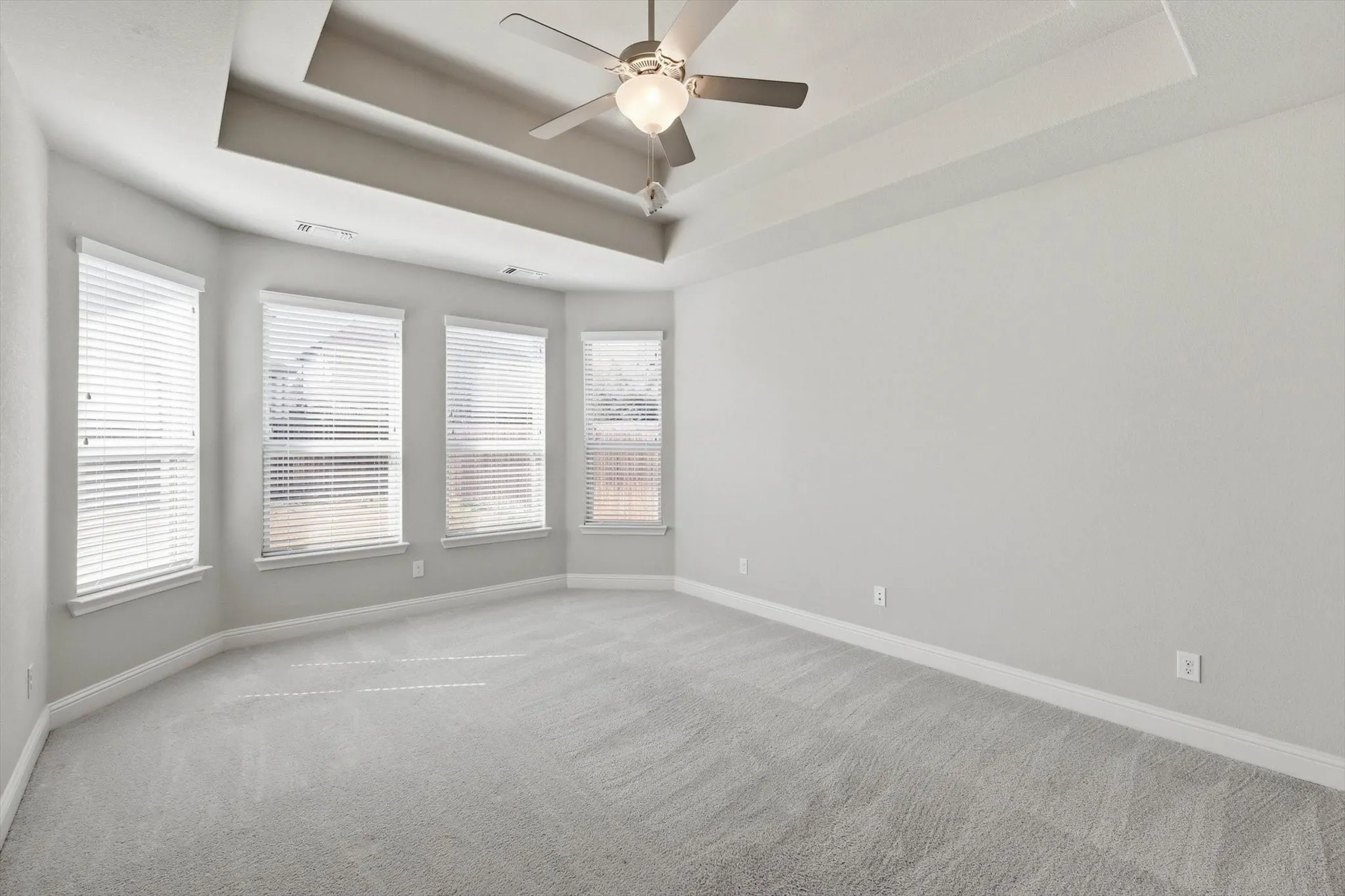 Spacious room featuring a tray ceiling, ceiling fan with light fixture, light gray carpeting, and multiple windows with blinds