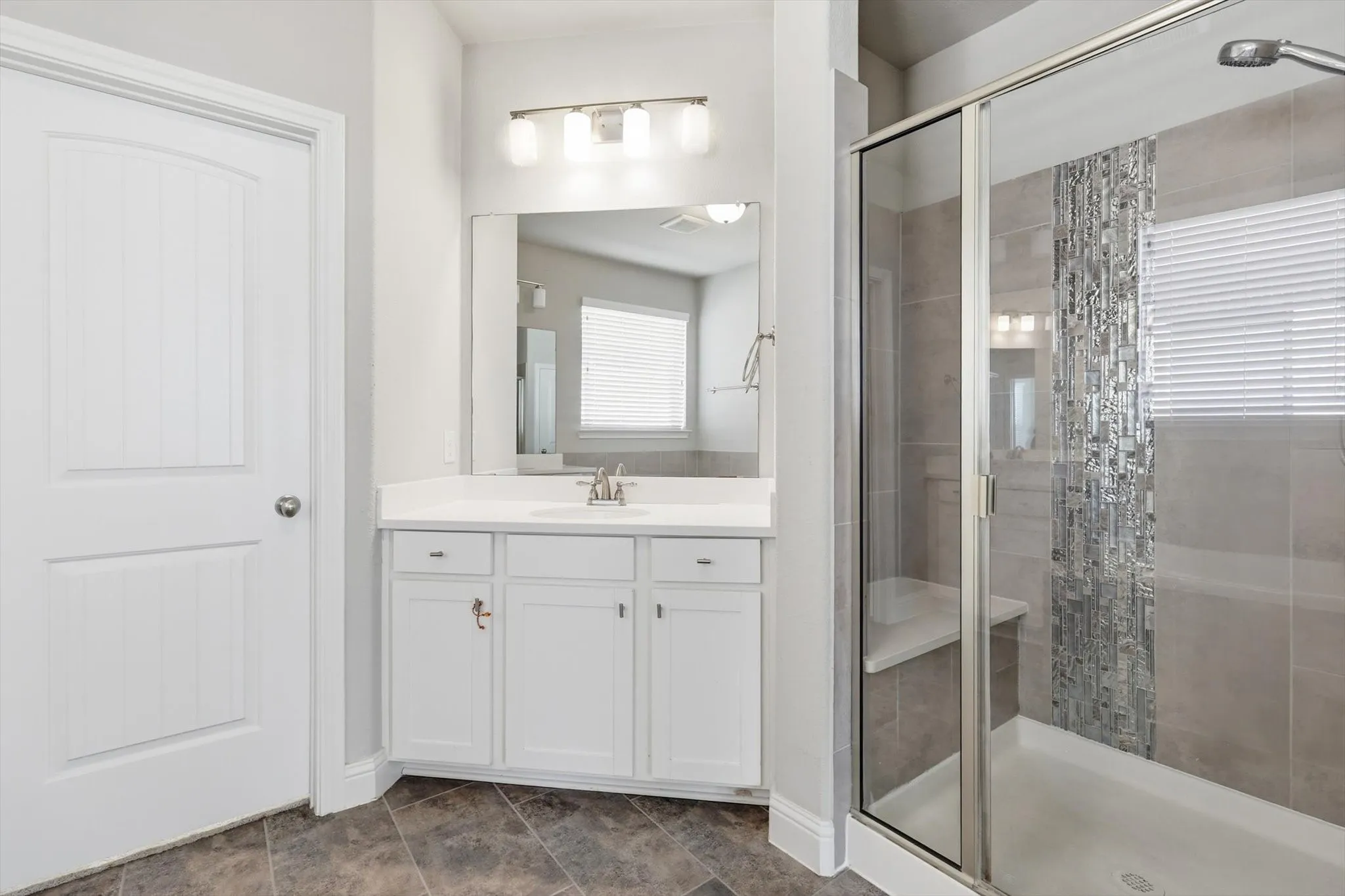 Bathroom featuring a single vanity with white cabinetry, a framed mirror, a three-light vanity fixture, a glass-enclosed shower with a built-in bench, and decorative tile accents