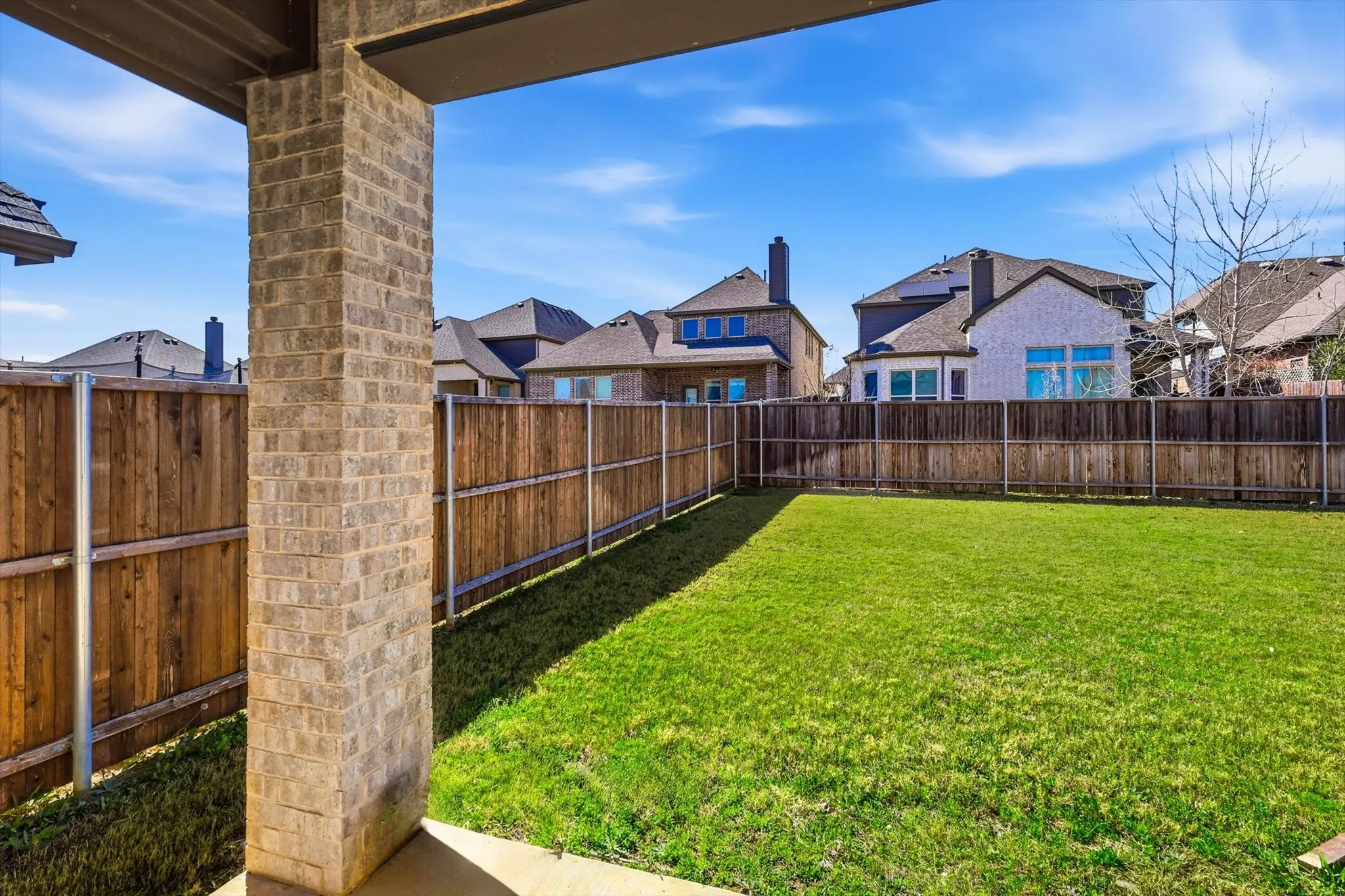 Expansive backyard featuring a manicured lawn, wood privacy fencing, and a covered patio with brick columns