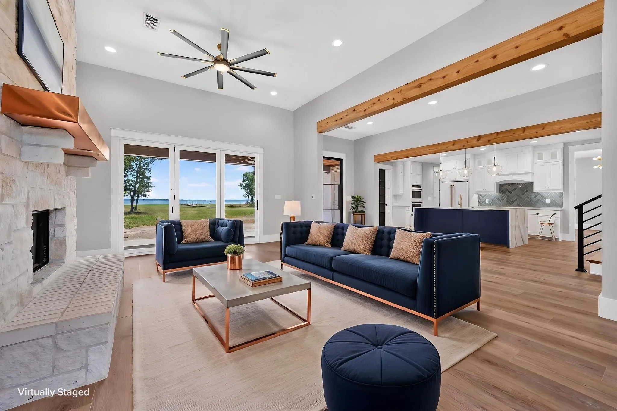 Magnificent open concept living room highlighted by soaring ceilings, timber accent beams, a striking stone fireplace with custom timber mantle, and an expansive view of the lake.