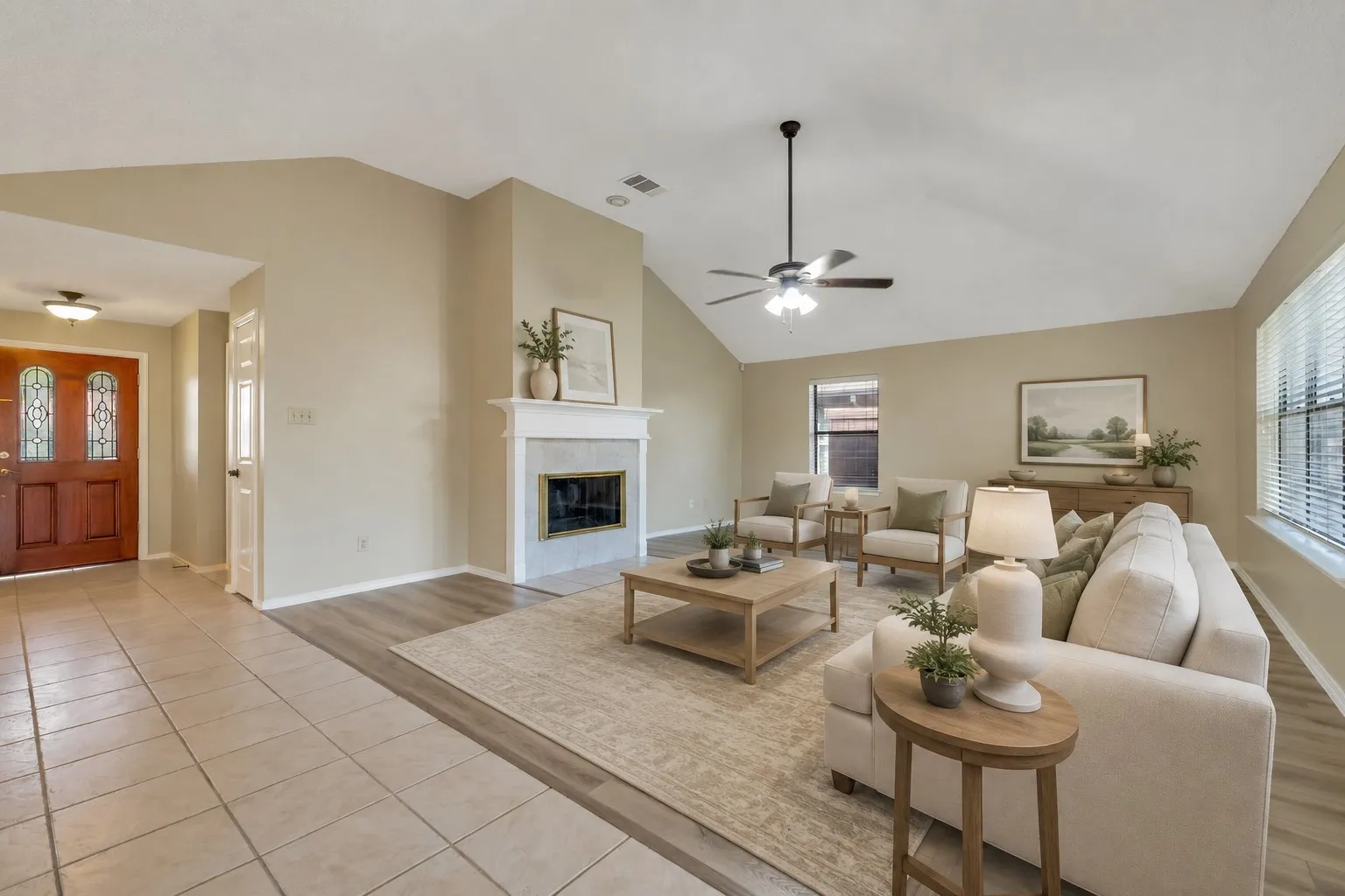Spacious living area featuring a vaulted ceiling with a ceiling fan, a prominent fireplace with a white mantel, and wood-finish flooring -virtually staged to illustrate possible furniture placement.