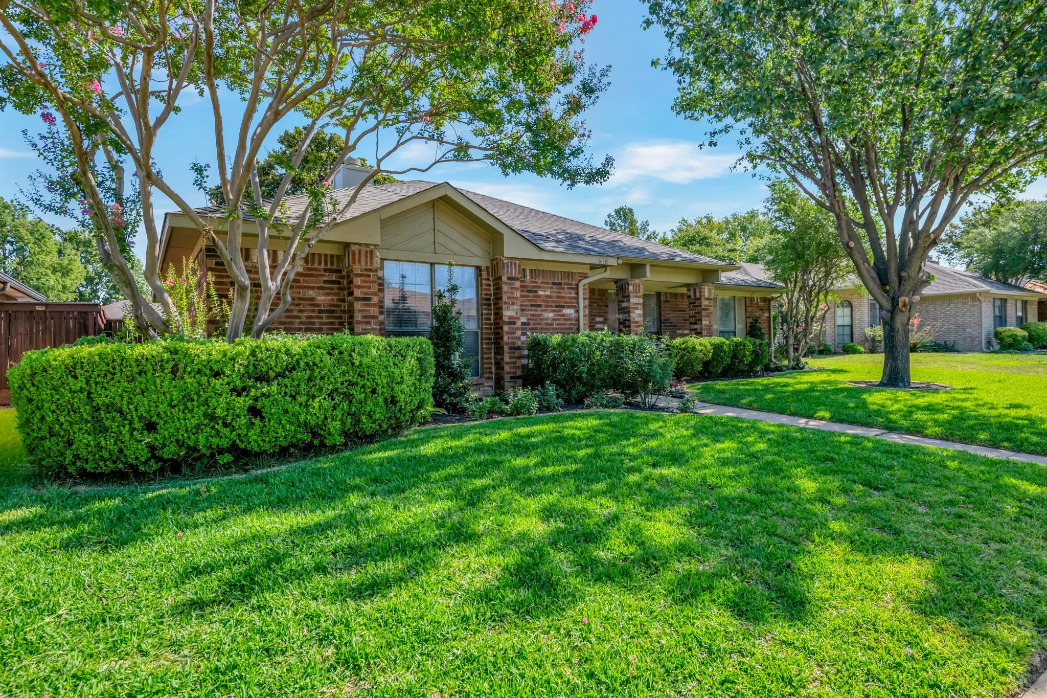 Brick exterior with a gabled roofline, mature trees, manicured shrubs, and a concrete walkway