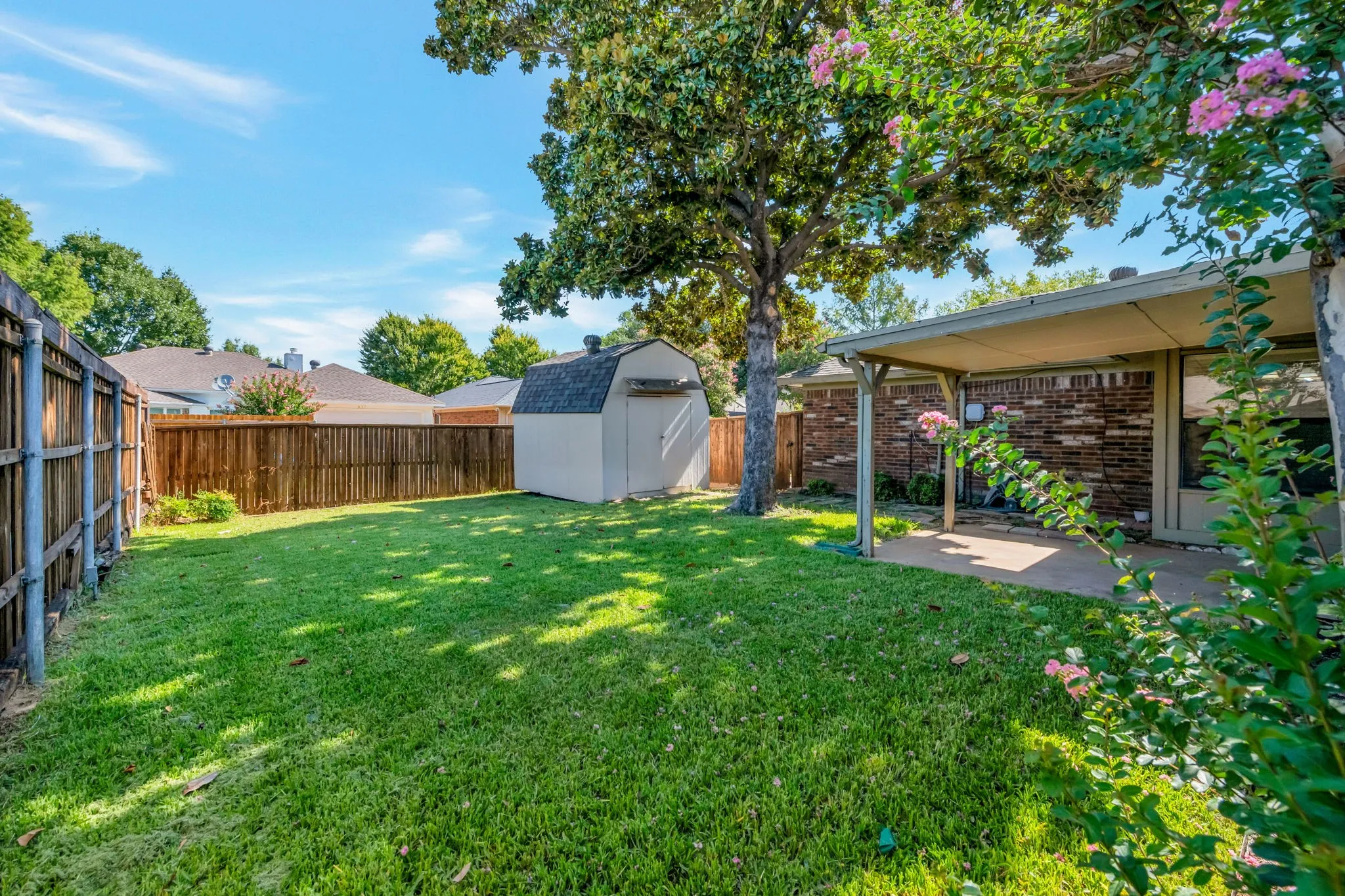 Expansive backyard featuring a covered patio, brick exterior, mature trees, wood fence, and an outdoor storage shed