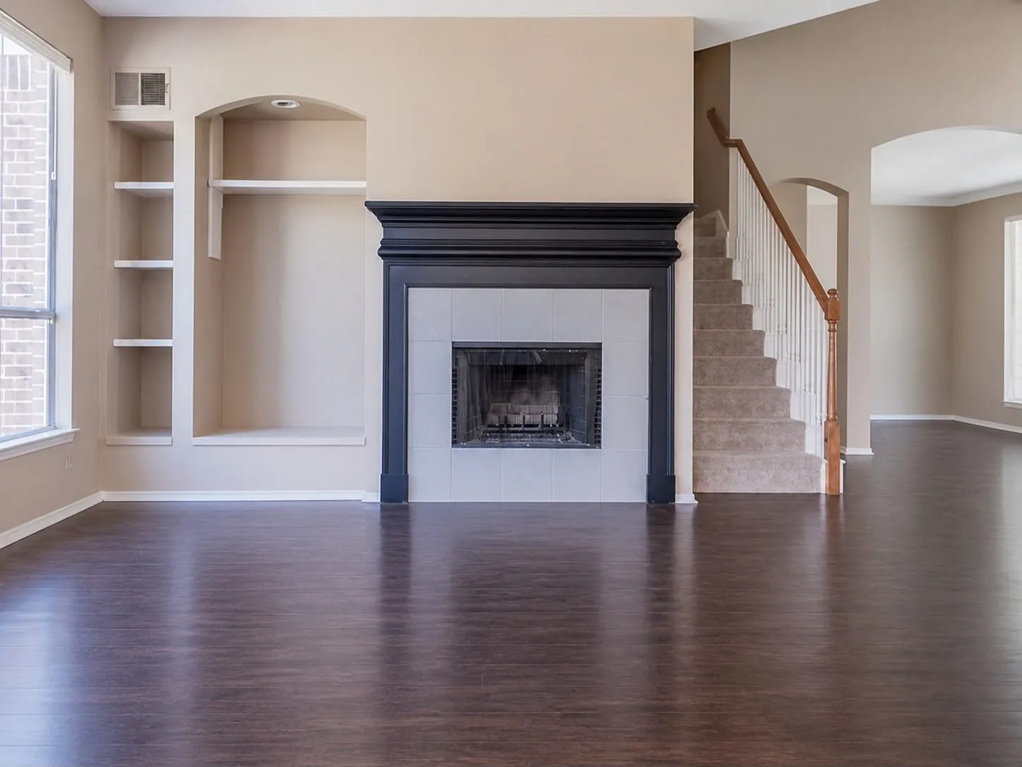 Living area featuring dark wood-finish flooring, a fireplace with a black mantel and white tile surround, built-in shelving, an arched alcove, and a carpeted staircase with a wood handrail
