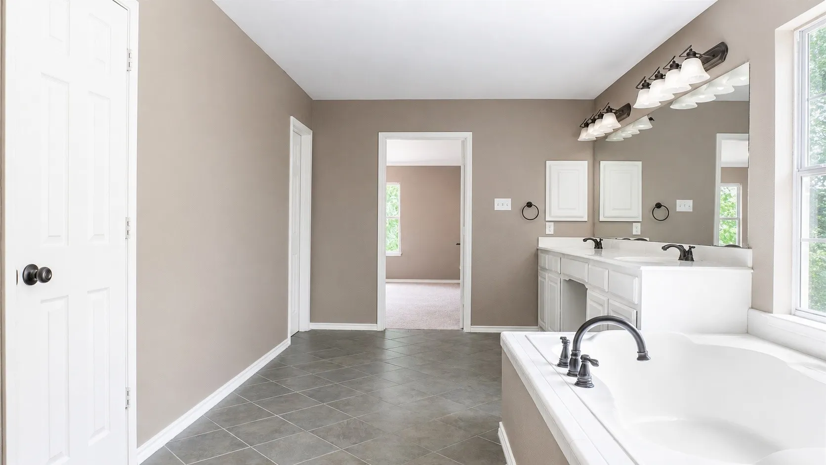 Bathroom featuring a soaking tub with a dark bronze faucet, dual vanity with white cabinetry, a large wall-mounted mirror, and tile flooring