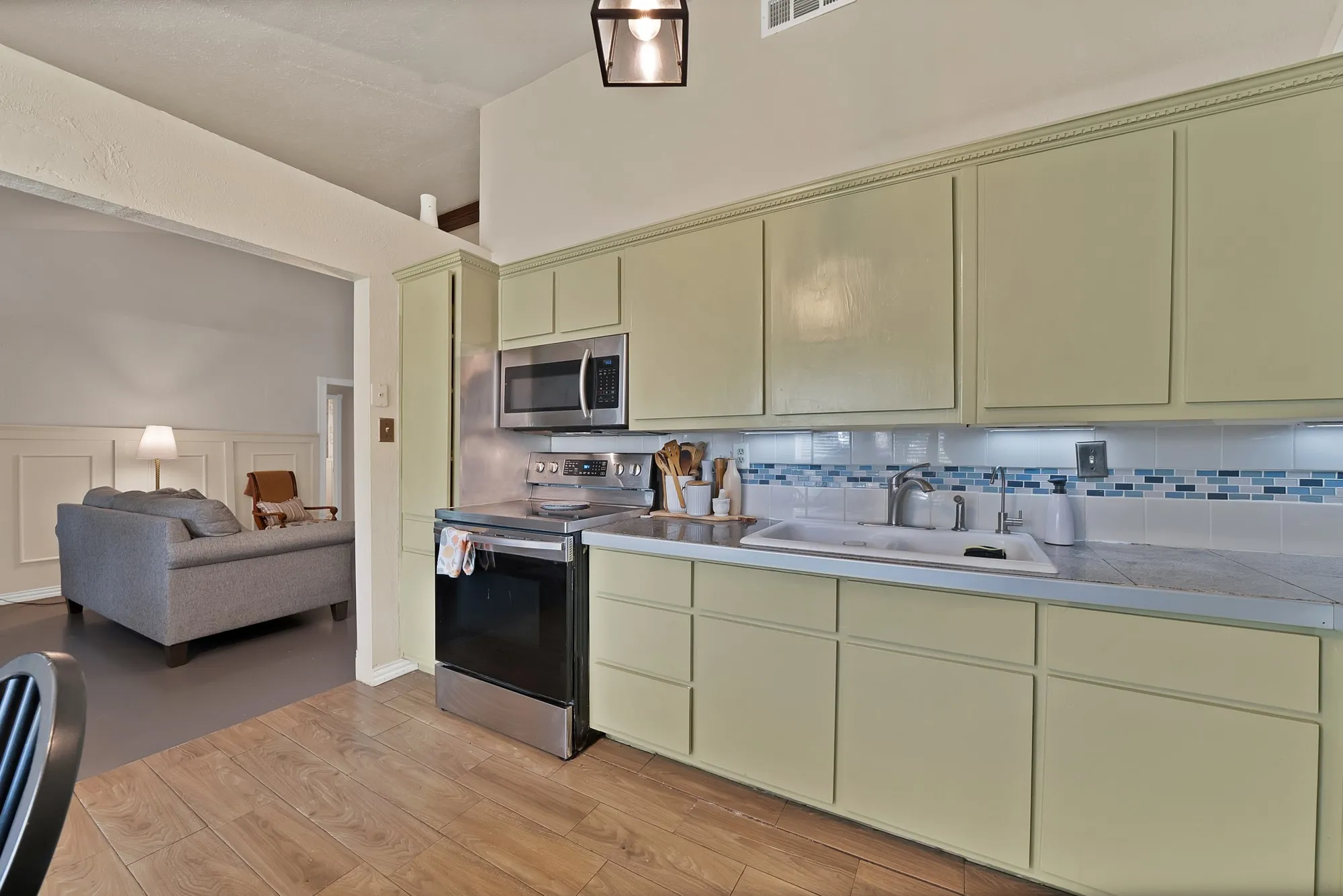 Kitchen featuring light green cabinetry, stainless steel appliances, wood-finish flooring, undermount sink, and a tile backsplash with mosaic accents