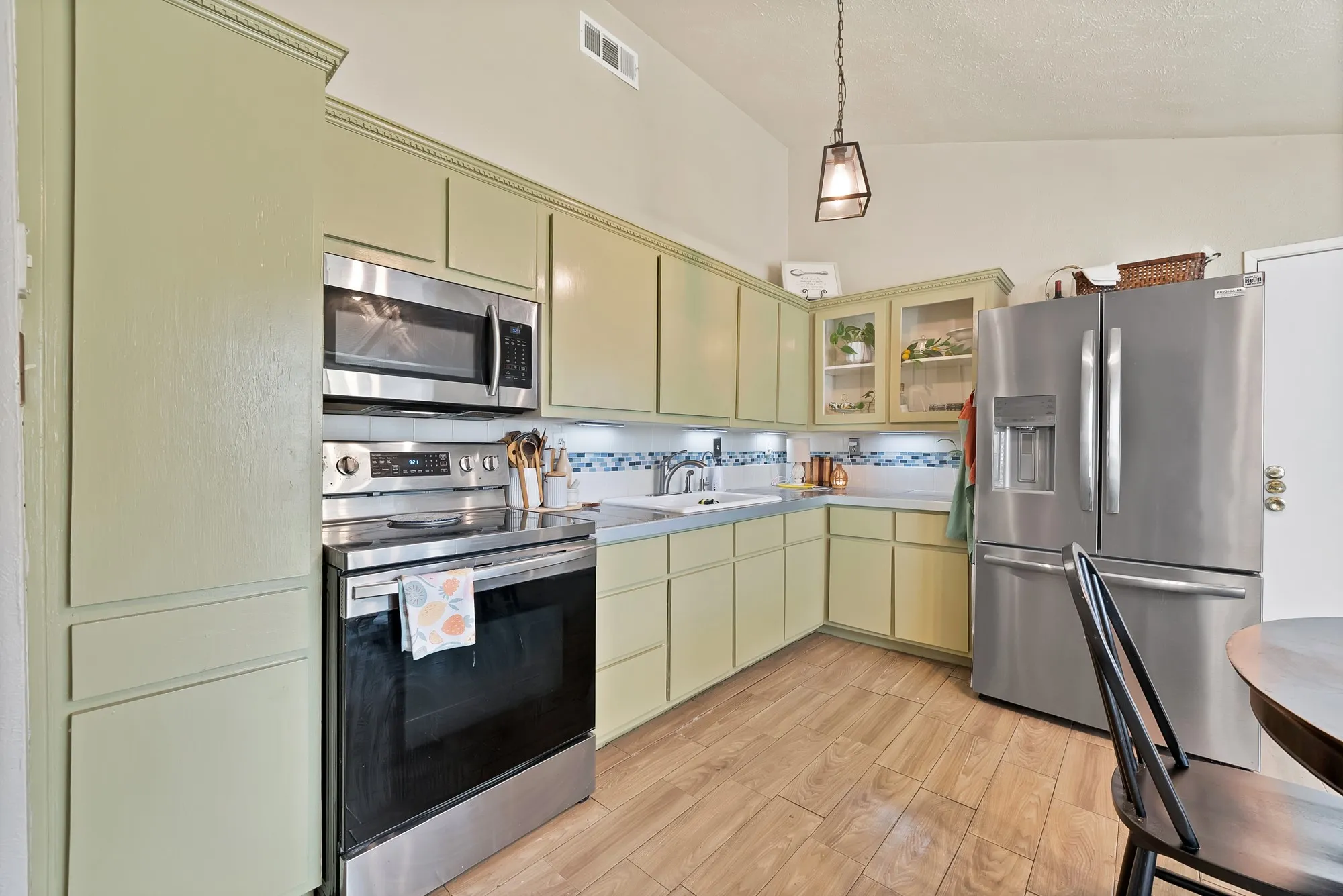 Kitchen featuring light green cabinetry, stainless steel appliances, wood-finish flooring, and a tiled backsplash