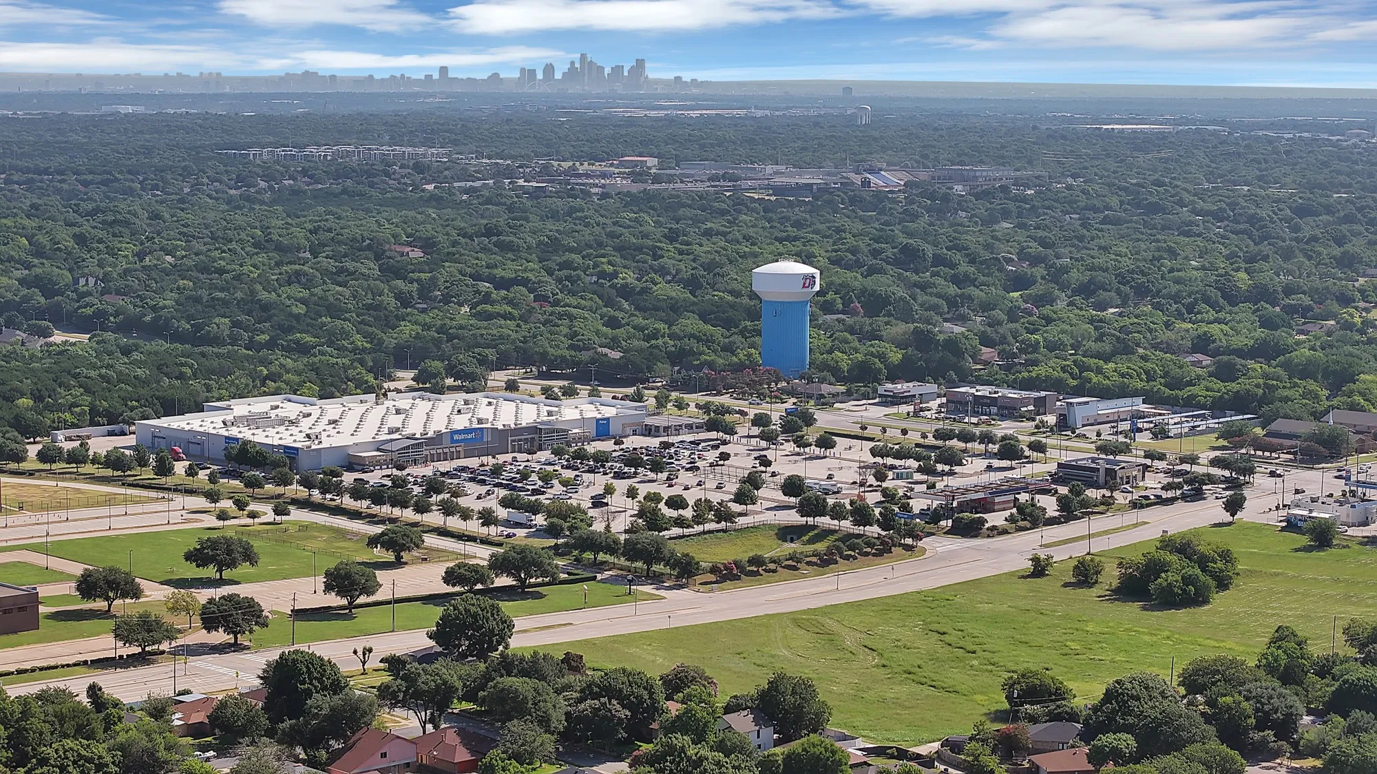 Expansive aerial highlighting a commercial district with retail storefronts, a large parking area, and surrounding green spaces
