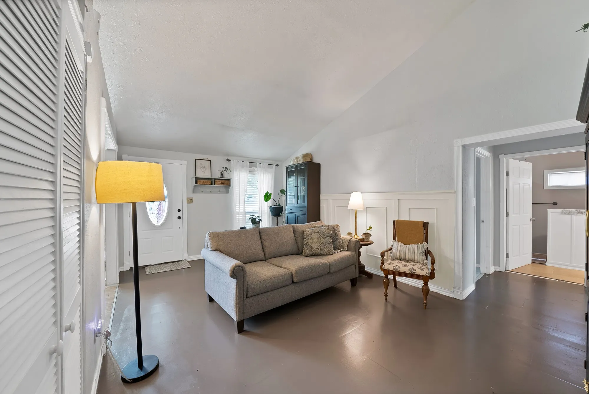 Living area with vaulted ceiling, white paneled accent wall, dark wide plank flooring, and a white paneled entry door