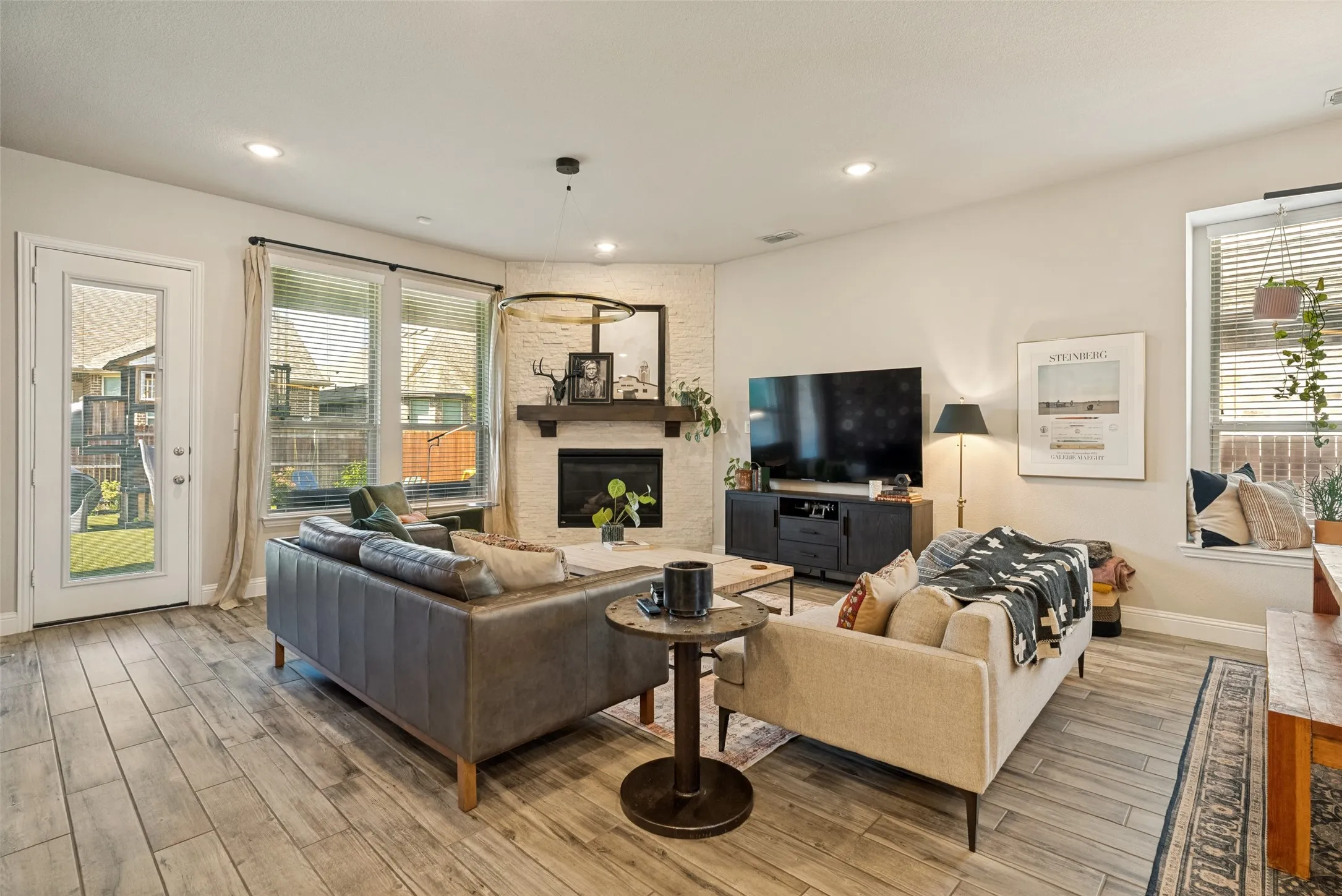 Living area featuring wood-finish flooring, a corner fireplace with a white stone surround and wood mantel, recessed lighting, and large windows with blinds
