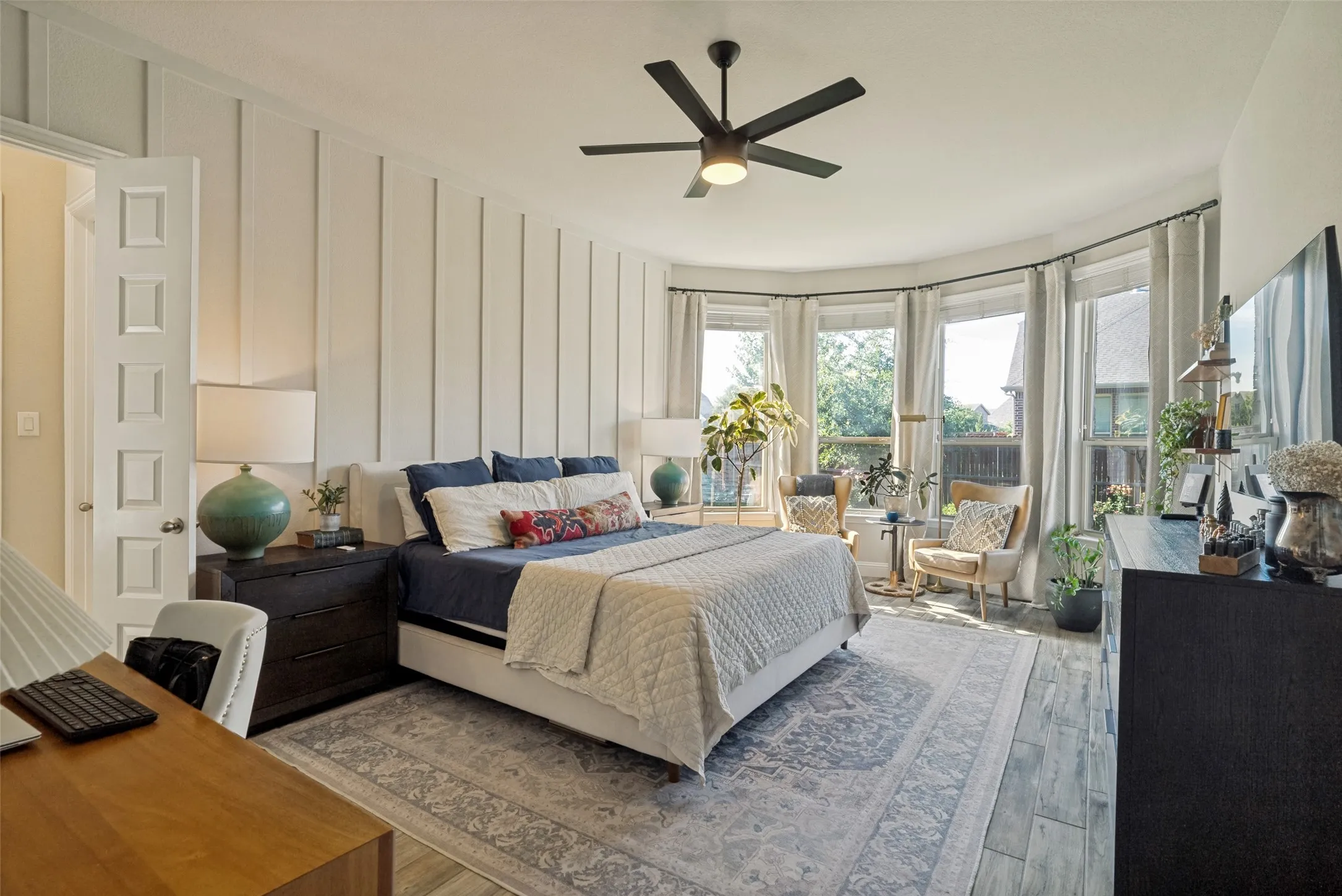 Spacious bedroom featuring a bay window, wood-finish flooring, a contemporary ceiling fan, and board and batten accent wall