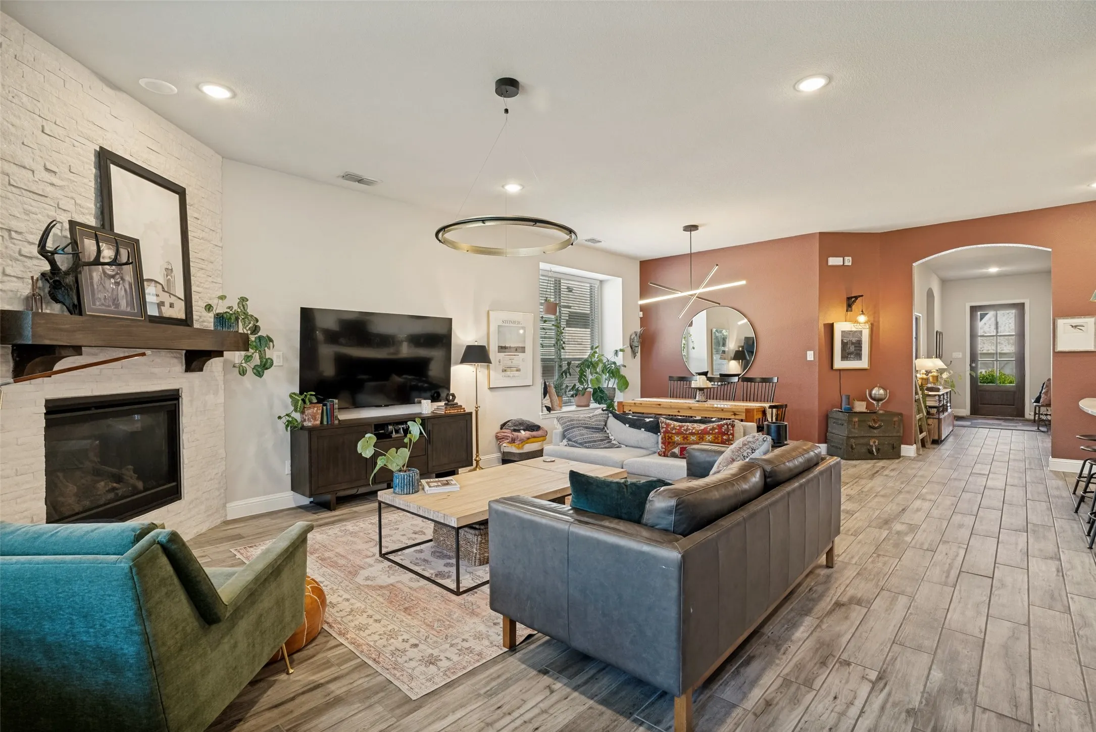 Spacious open-concept living area featuring wood-finish tile flooring, a fireplace with a white stone surround and wood mantel, recessed lighting, and an arched entryway