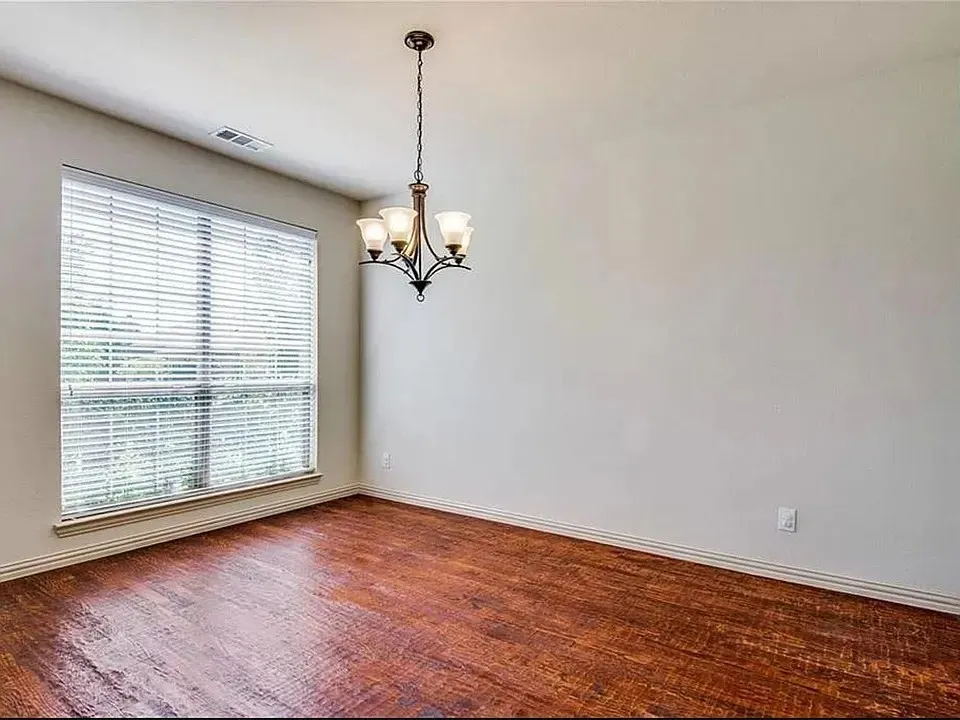 Spacious room featuring wood-finish flooring, a large window with blinds, and a multi-arm chandelier