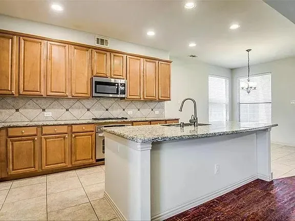 Kitchen featuring a central island with an integrated sink, granite countertops, wood cabinetry, stainless steel appliances, and tile backsplash