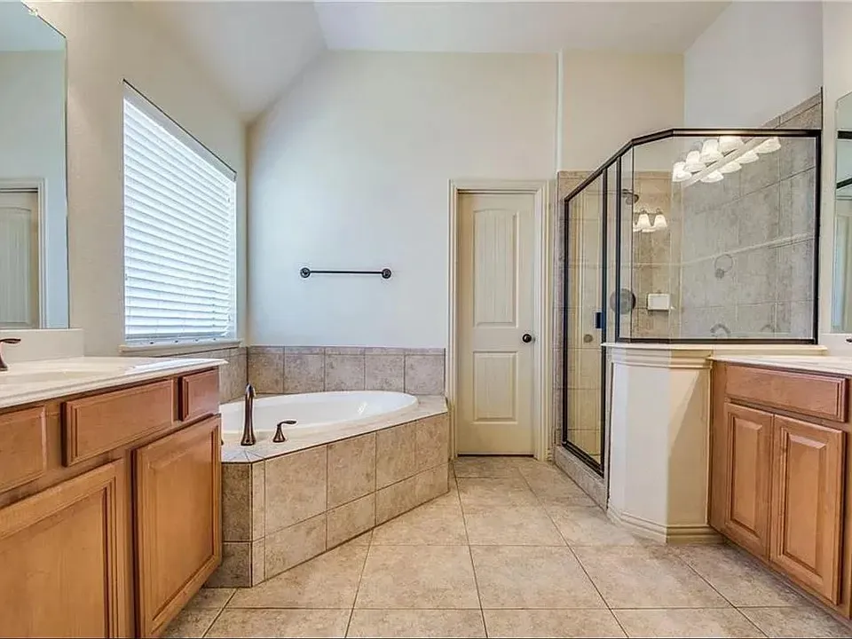 Bathroom featuring a corner soaking tub with tiled surround, a separate shower with a glass enclosure, dual vanity sinks with light wood cabinetry and countertops, and tiled flooring