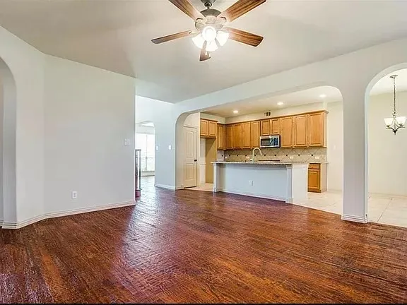 Spacious interior with wood-finish flooring, arched doorways, a ceiling fan, and an integrated kitchen featuring wood cabinetry, stainless steel appliances, and a breakfast bar with a stone-look countertop