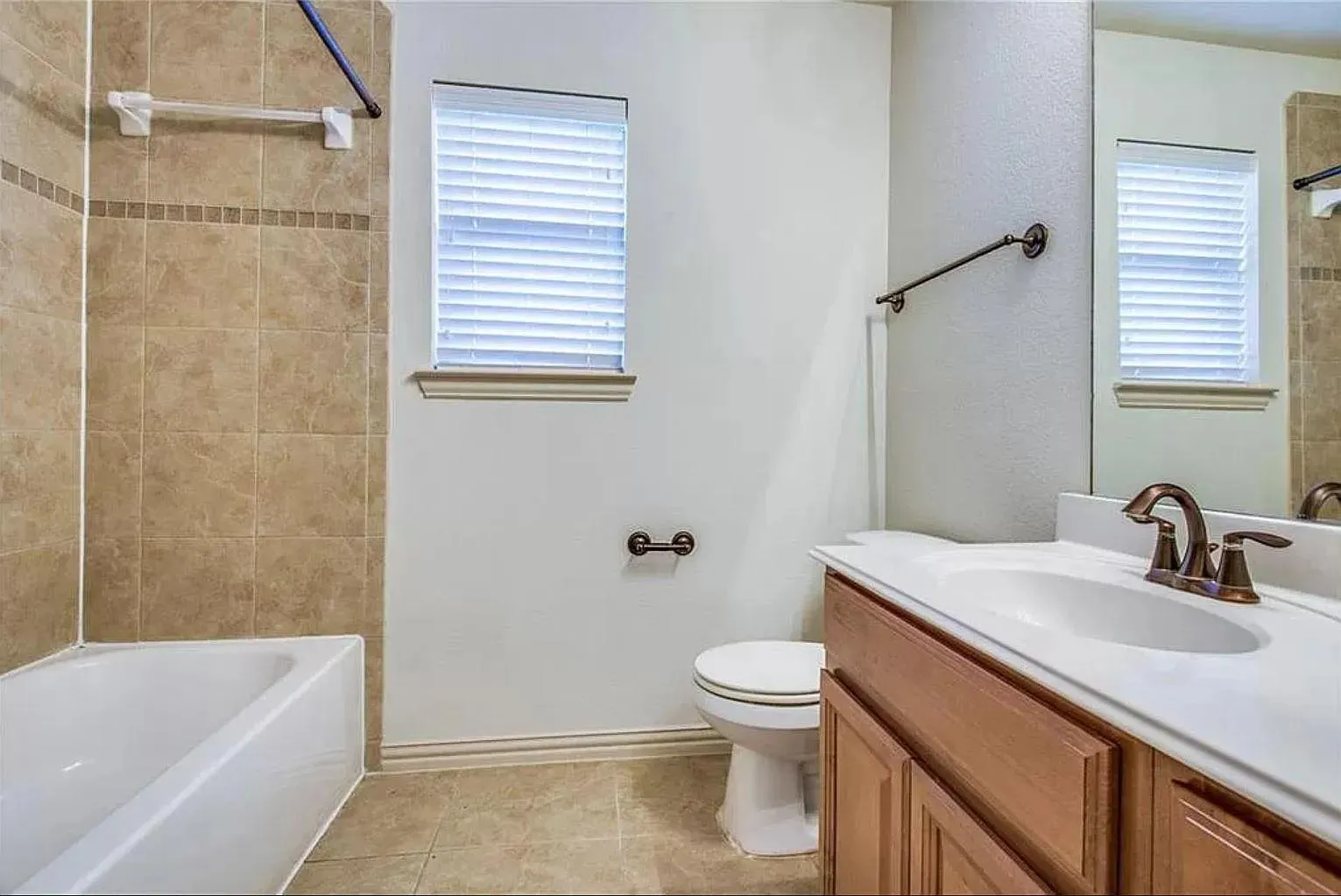 Bathroom featuring a white bathtub, floor-to-ceiling tiled shower surround with accent band, single window with blinds, and a vanity with an integrated sink and bronze-finish faucet