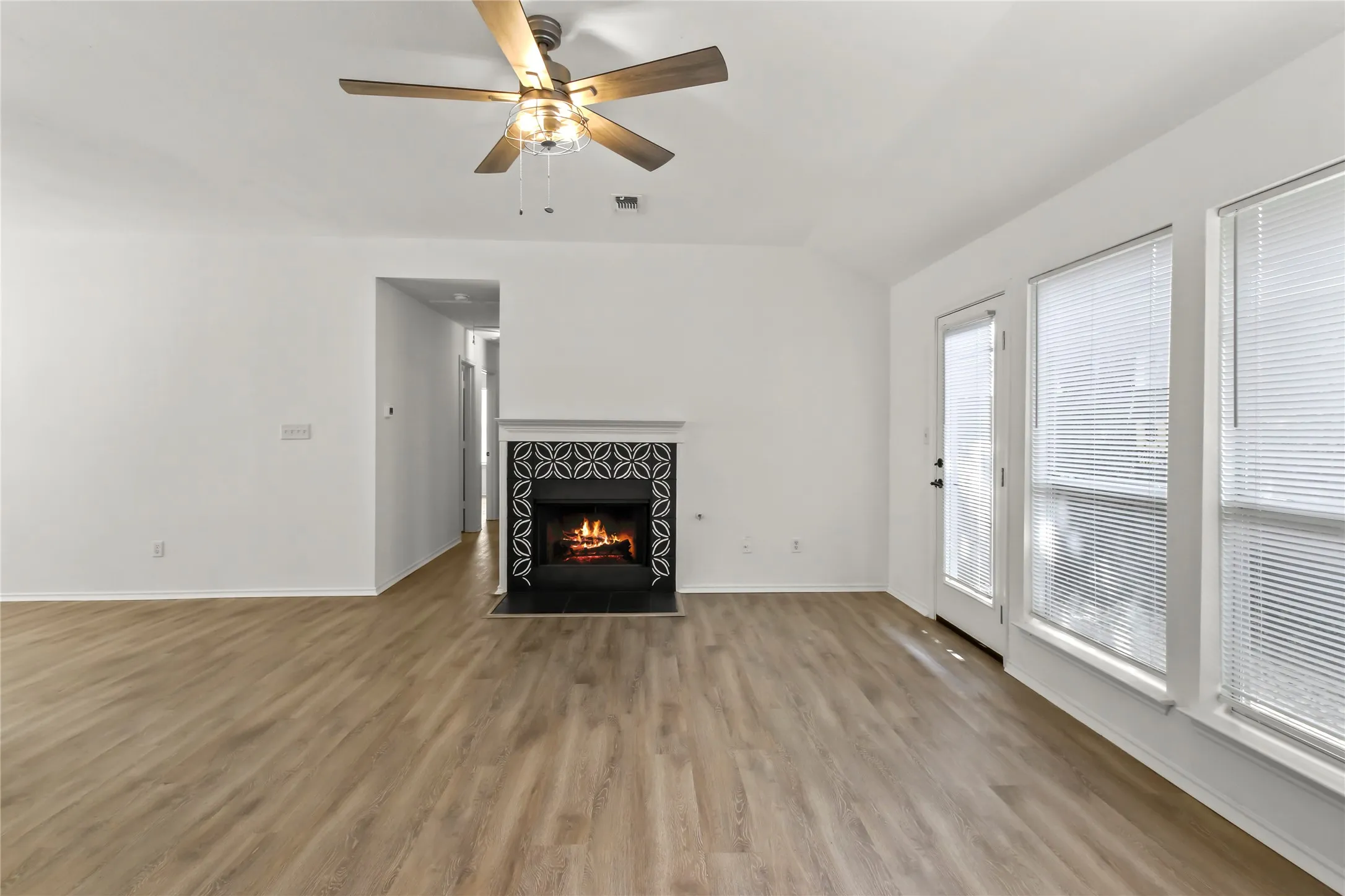 Spacious living area featuring a decorative tile fireplace with white mantel, wood-finish flooring, and multiple windows with blinds