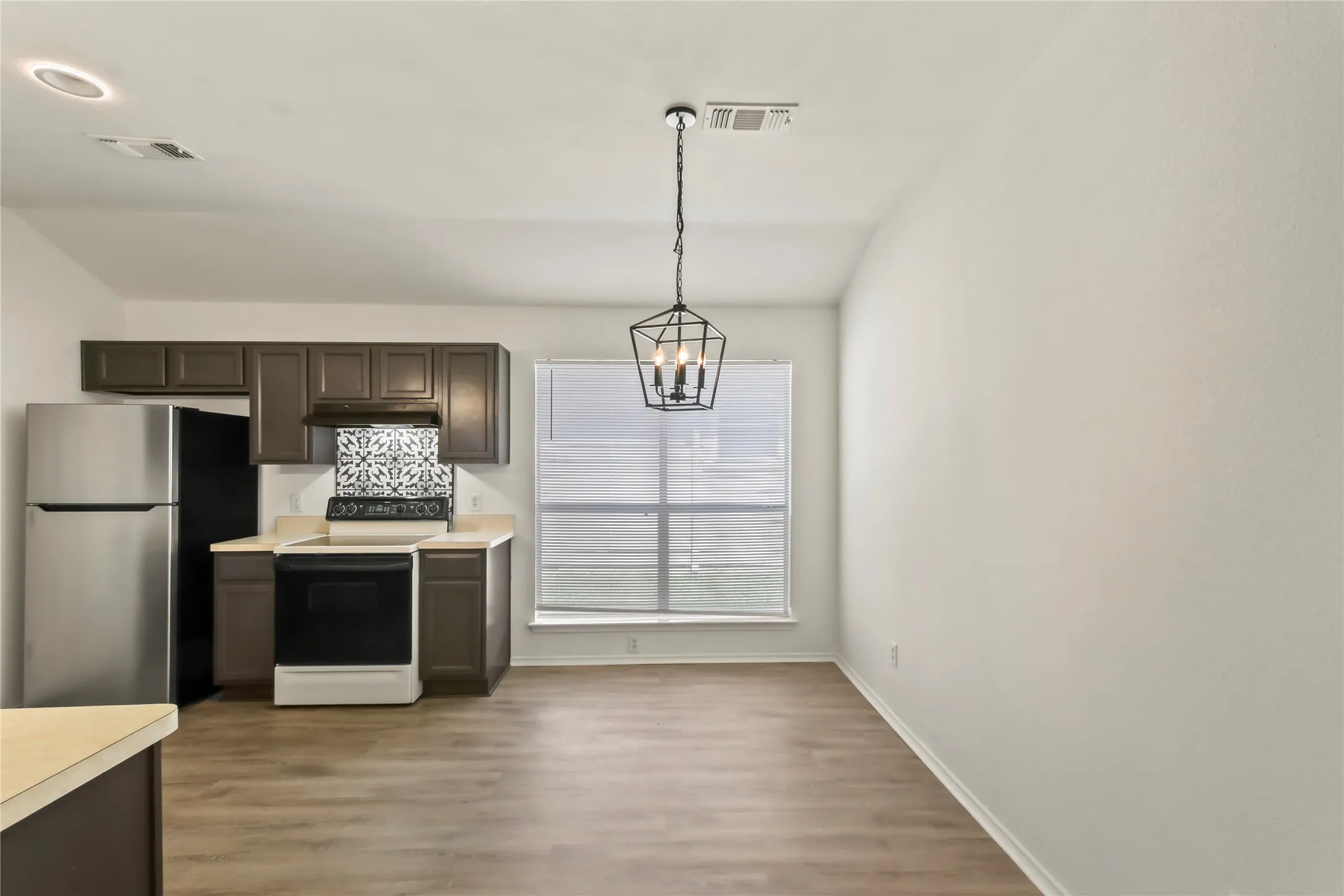 Open-concept kitchen and dining area featuring wood-finish flooring, dark wood cabinetry, stainless steel appliances, and a patterned tile backsplash