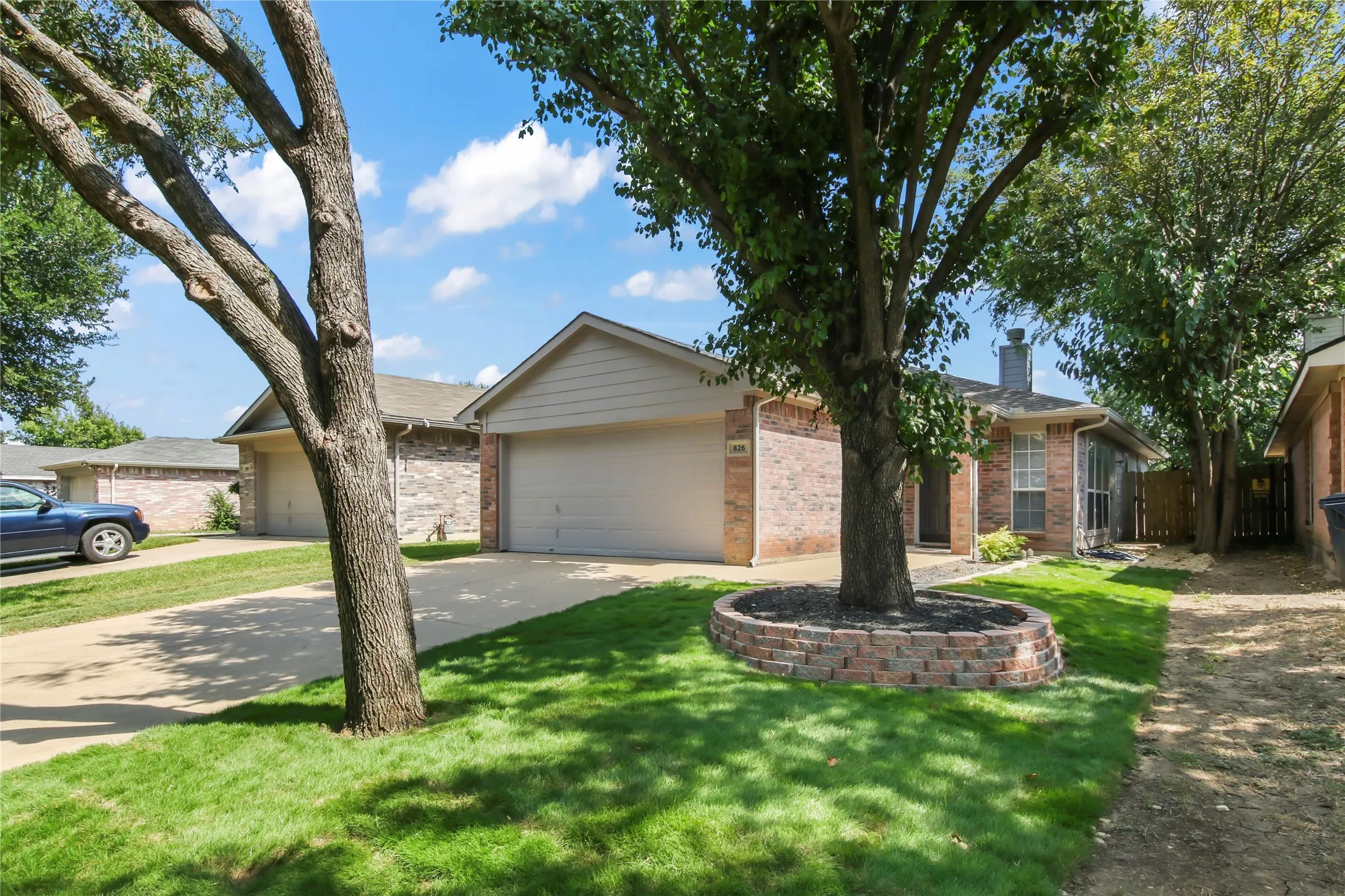 Brick exterior featuring an attached two-car garage, mature trees, a manicured lawn, and a decorative stone-lined planting bed