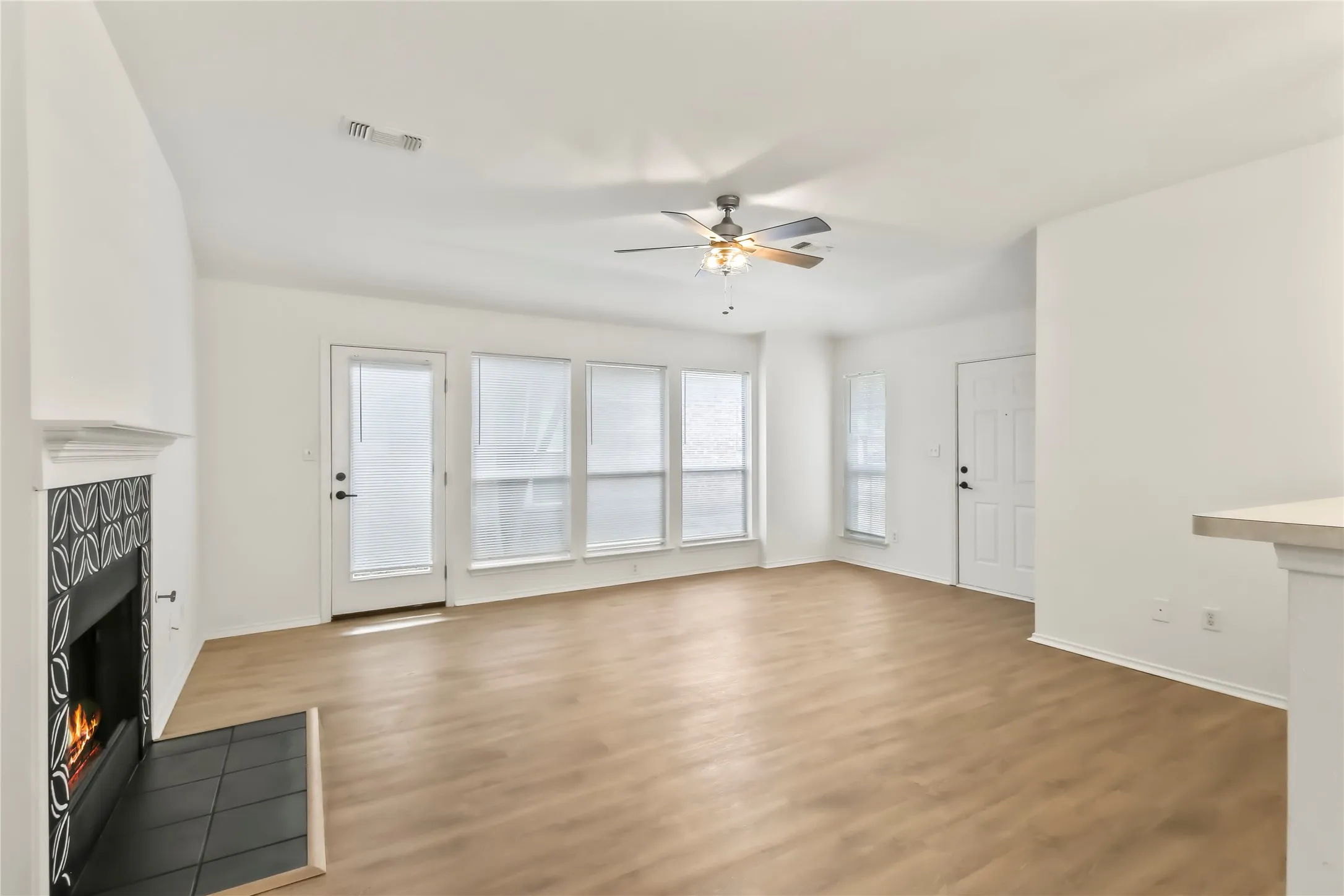 Spacious living area featuring wood-finish flooring, a decorative fireplace with patterned tile surround, multiple windows with blinds, a ceiling fan with integrated lighting, and a white paneled door