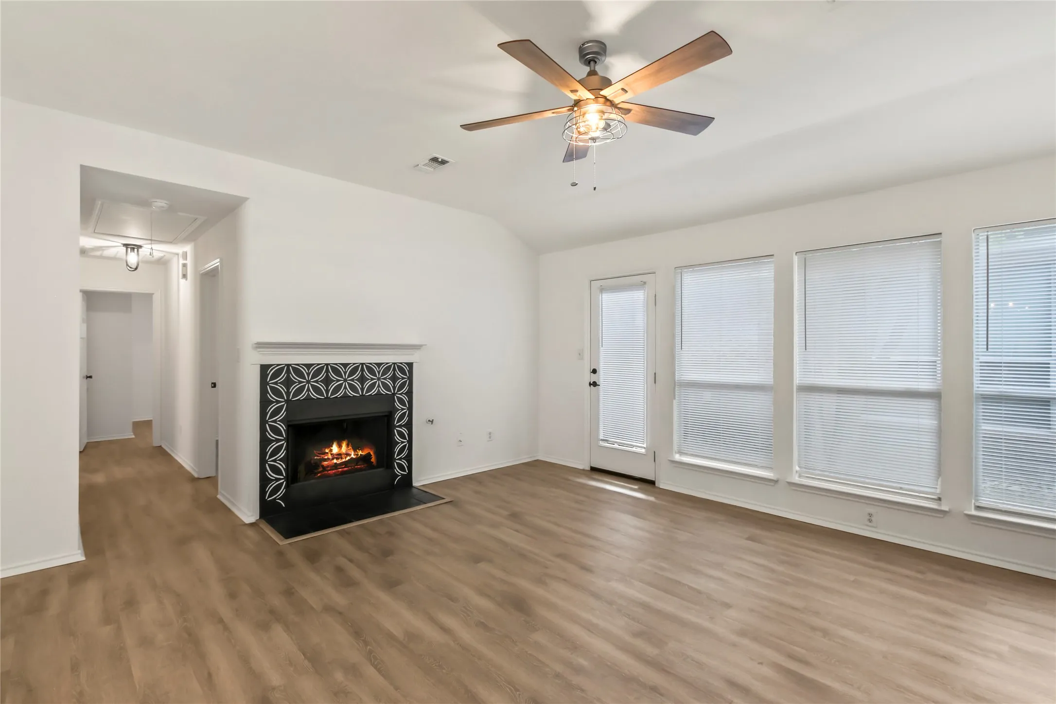 Living area featuring wood-finish flooring, a fireplace with decorative tile surround and black hearth, and multiple windows with blinds