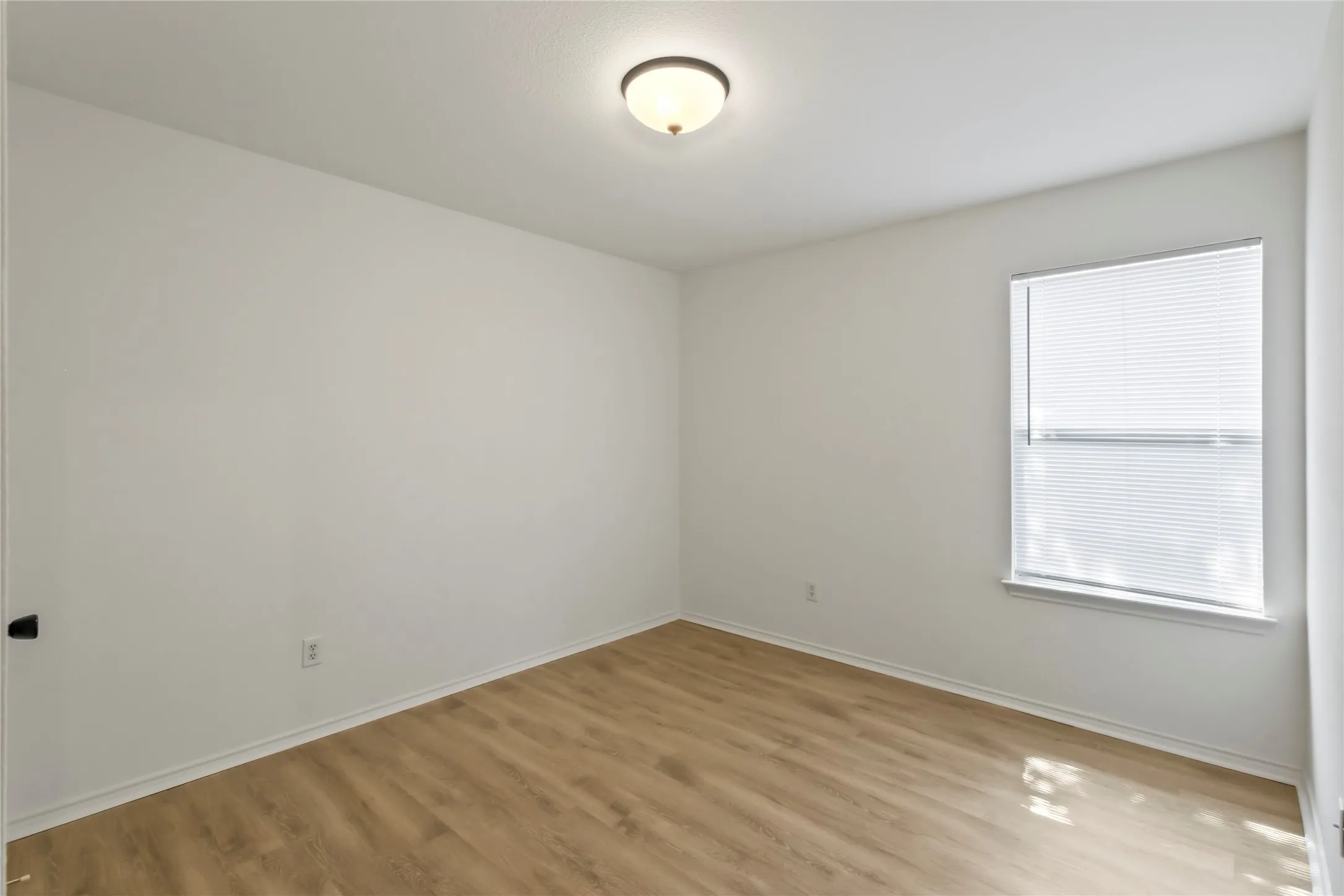 Bright interior space featuring light wood-finish flooring, a single window with blinds, and white painted walls