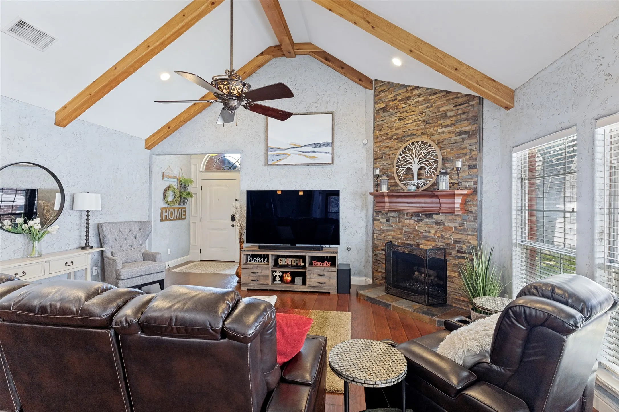 Vaulted ceiling with exposed wood beams, a prominent stacked stone fireplace, wood-finish flooring, an arched entryway, and multiple windows with blinds