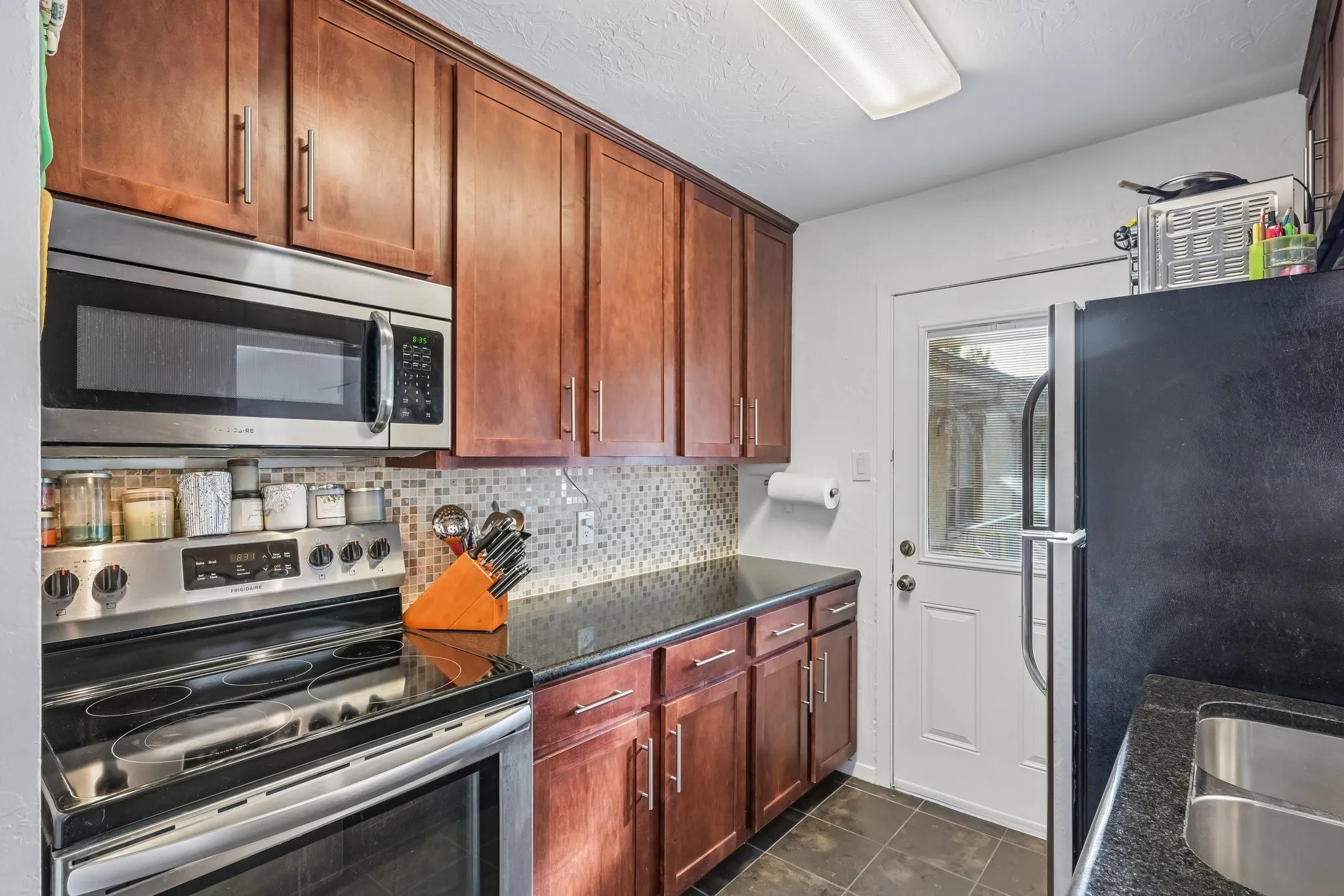 Kitchen featuring wood-finish cabinetry, black countertops, and a mosaic tile backsplash