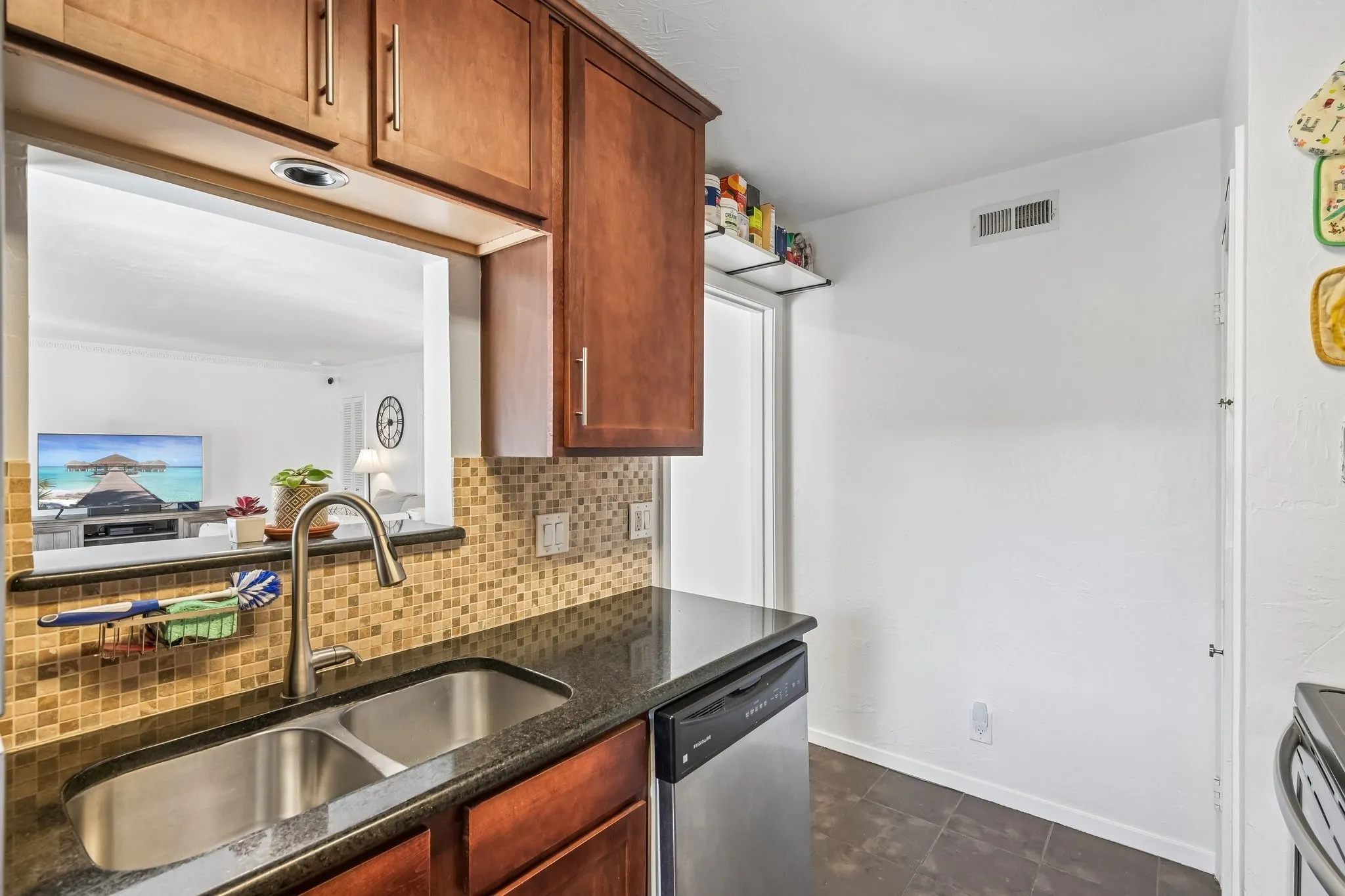 Kitchen featuring wood-finish cabinetry, a pass-through window, a stainless steel double basin sink with a gooseneck faucet, a mosaic tile backsplash, and dark countertop surfaces