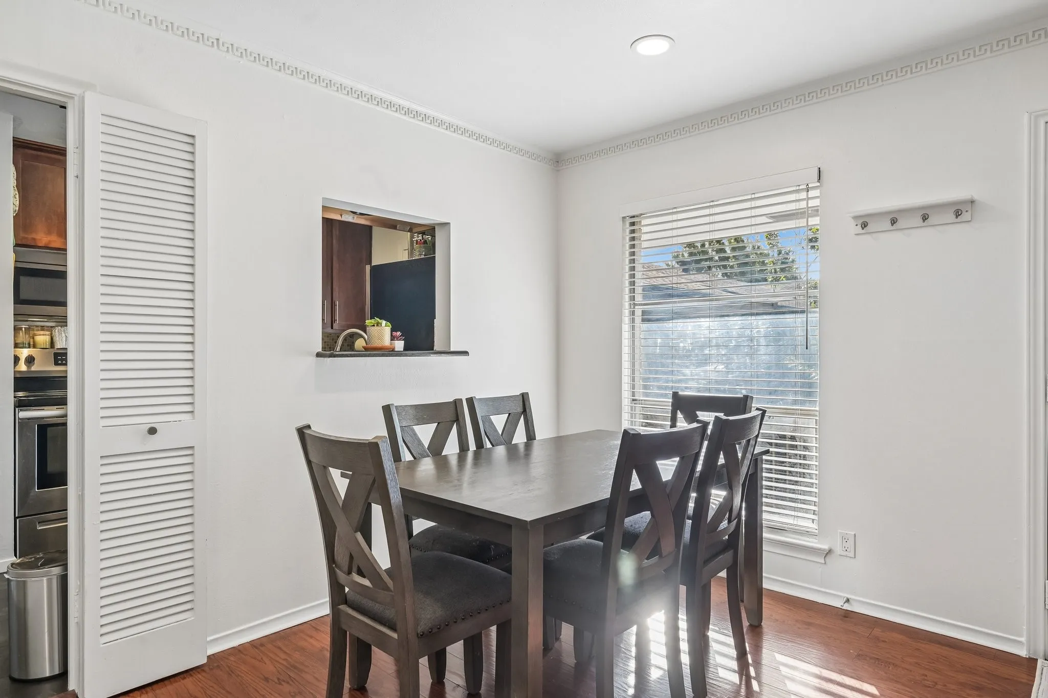 Dining area featuring hardwood flooring, a pass-through window, a recessed ceiling light, decorative crown molding, and a window with horizontal blinds