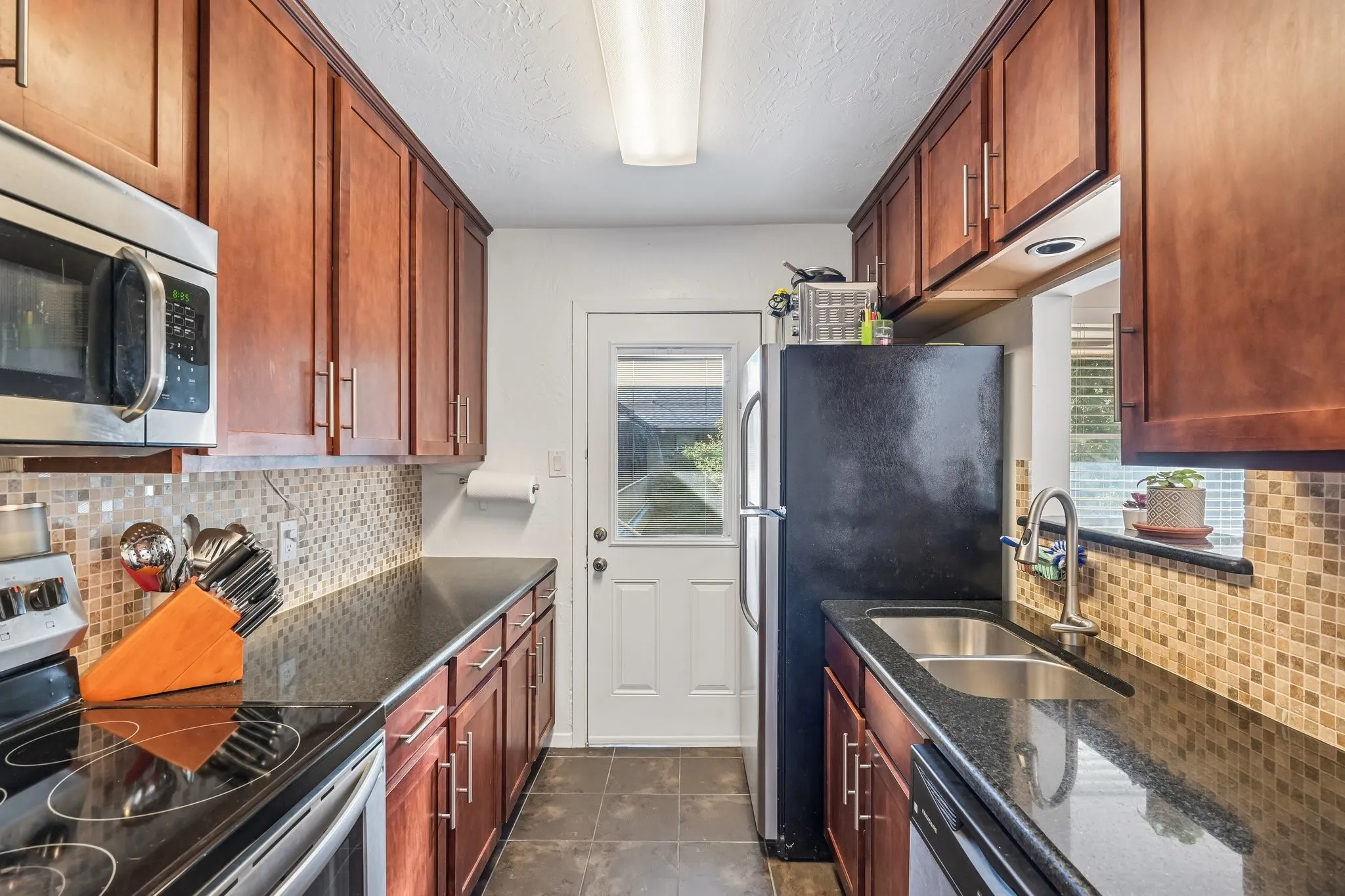 Galley kitchen featuring wood cabinetry, dark countertops, a mosaic tile backsplash, and stainless steel appliances including a built-in microwave and electric range
